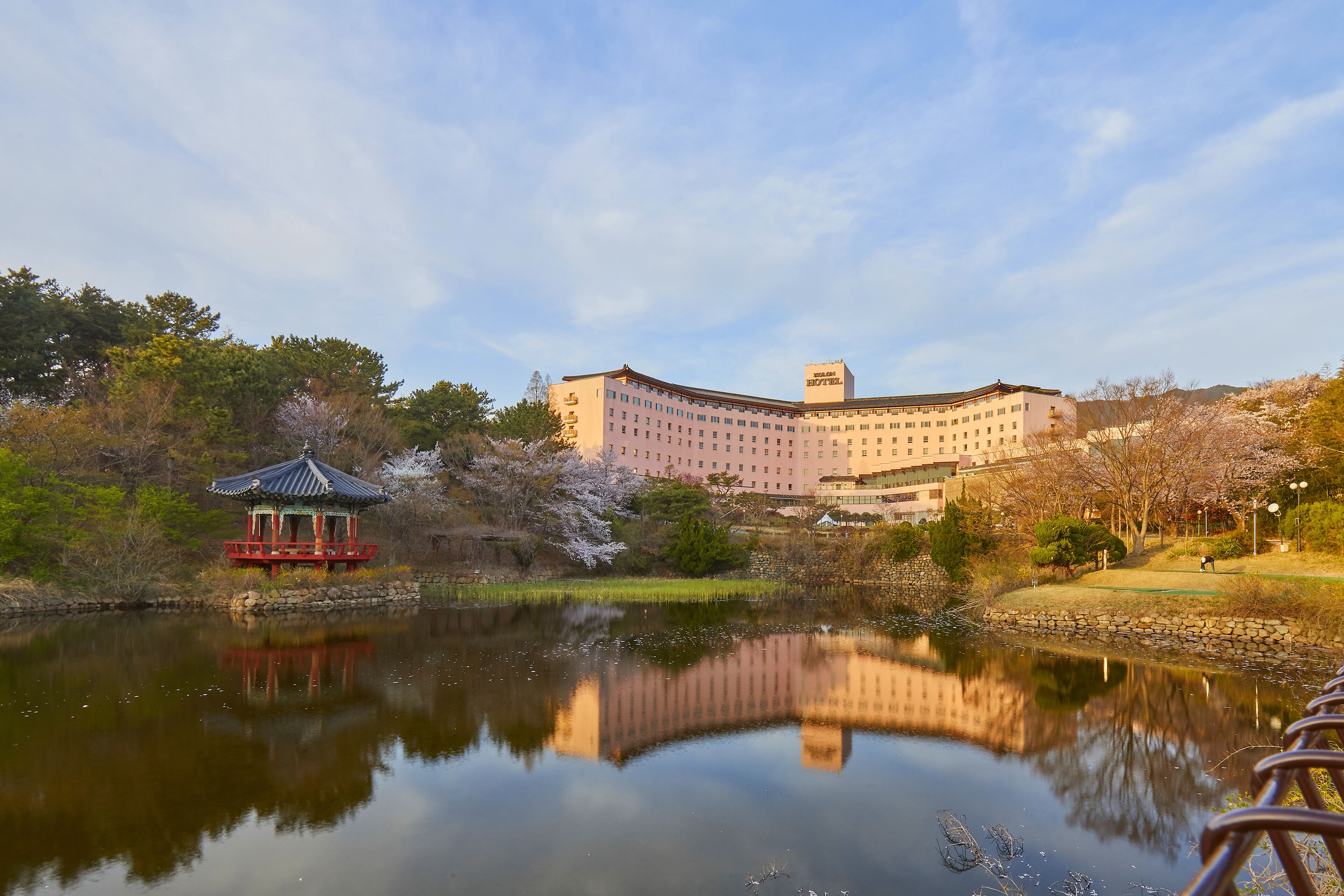 A large hotel with a reflection in a calm lake, surrounded by trees and a gazebo.
