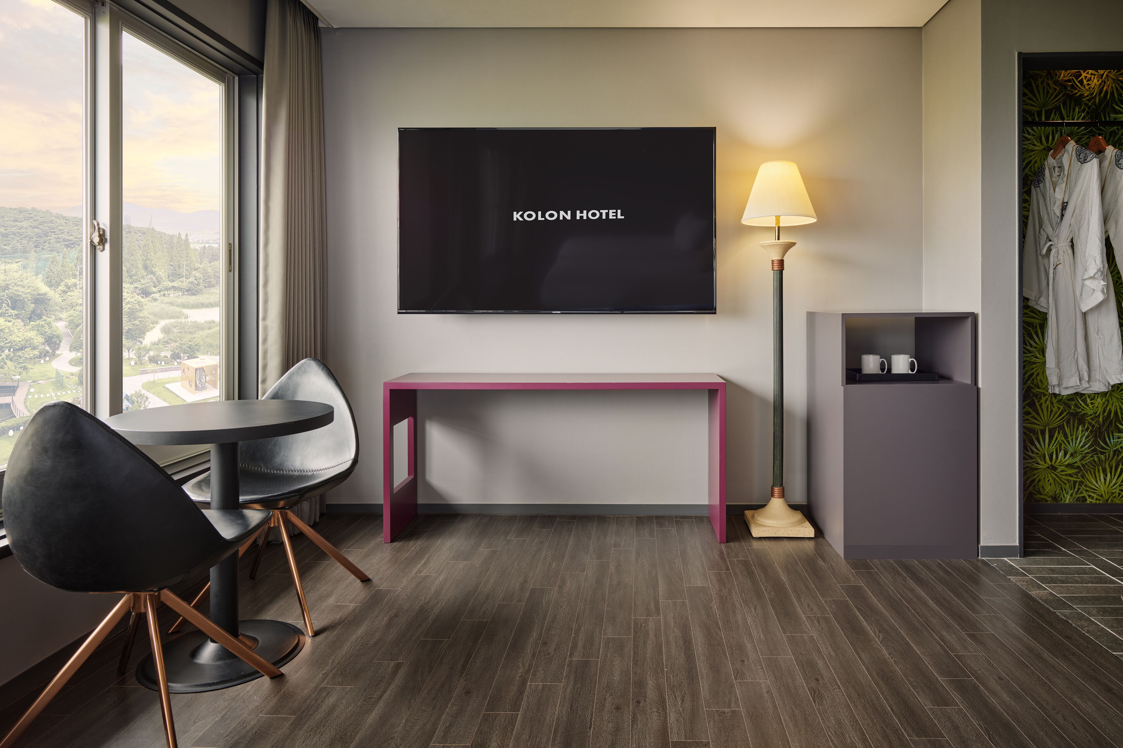 Habitación de hotel con un ventanal grande, un televisor de pantalla plana, un escritorio con una silla, una lámpara y un perchero para bata.