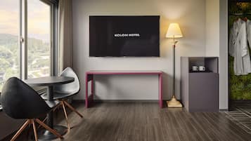 Habitación de hotel con un ventanal grande, un televisor de pantalla plana, un escritorio con una silla, una lámpara y un perchero para bata.