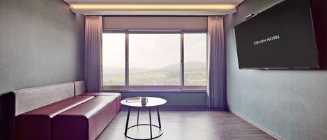 A modern hotel room with a large window, a sofa, a small round table, and a flat-screen TV on the wall.