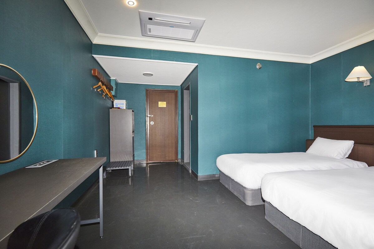 A hotel room with teal walls, two beds, a desk, a chair, a wardrobe, and a mirror.