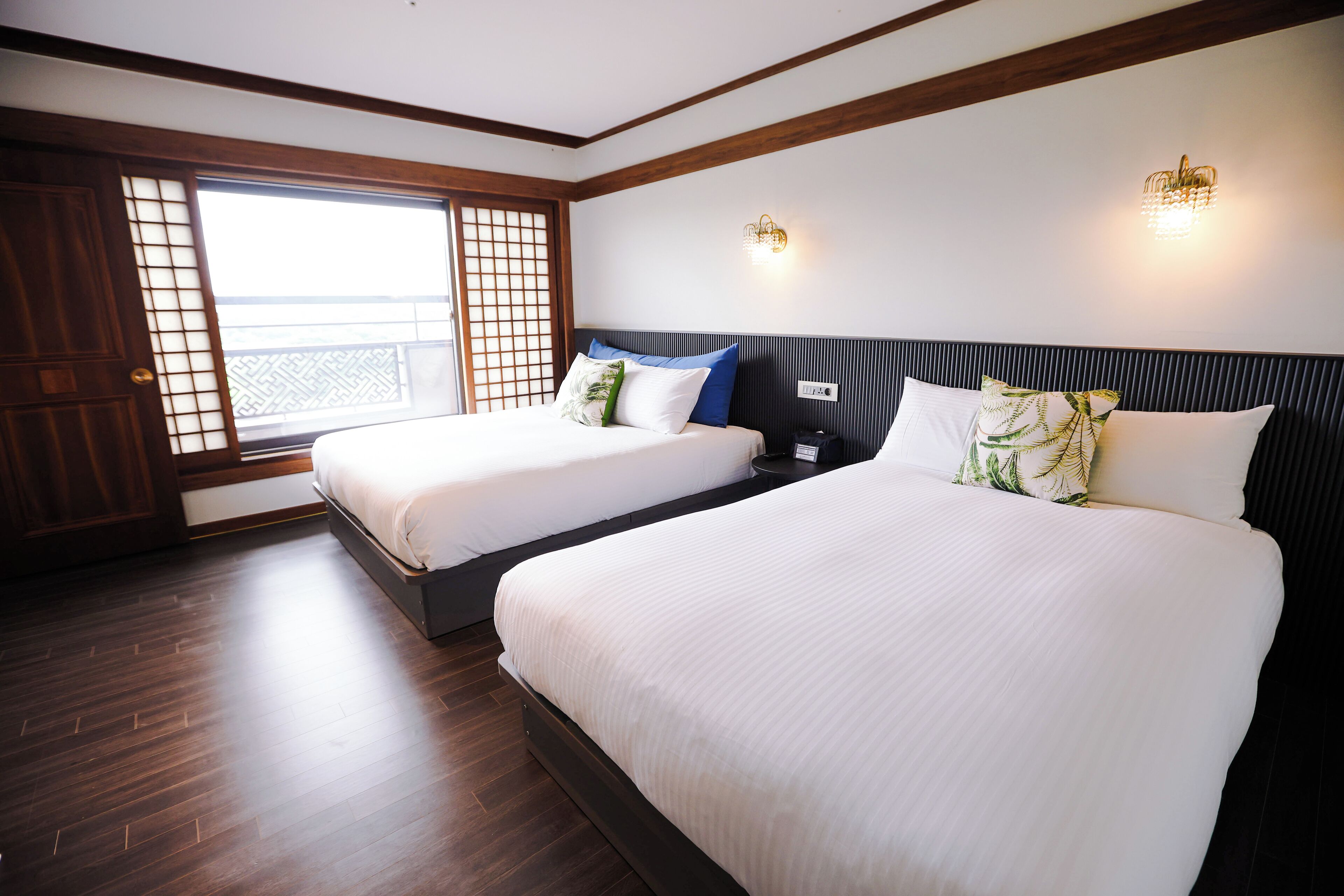 A hotel room with two beds, wooden flooring, a large window with a view, and wall-mounted lighting.