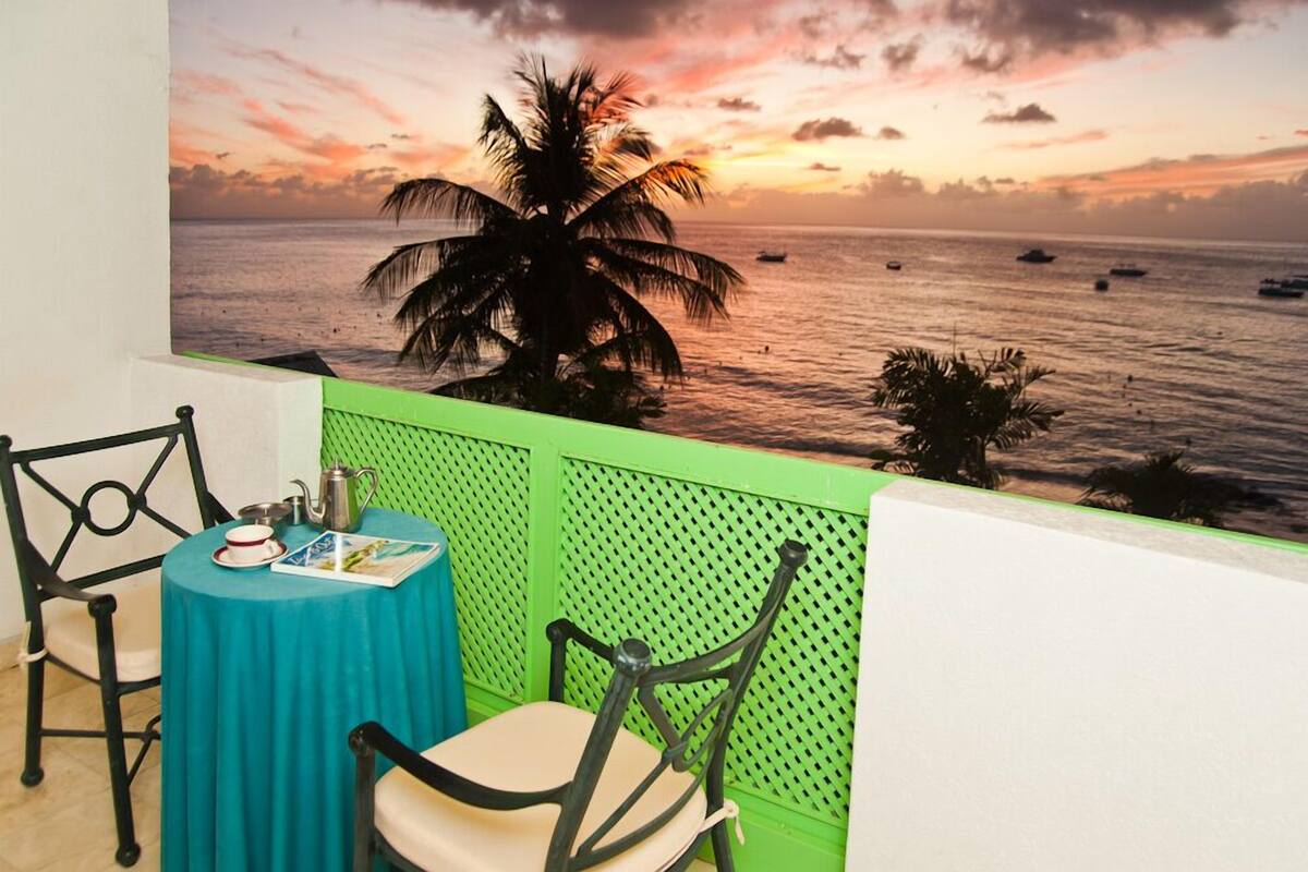A balcony with a view of the sea at sunset, a table with a green tablecloth, and two chairs.