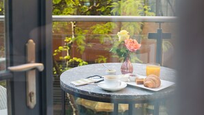Una mesa redonda con superficie de vidrio, un florero con flores, un croissant, un vaso de jugo de naranja, una taza de café y un teléfono inteligente en un balcón con vistas a la vegetación.