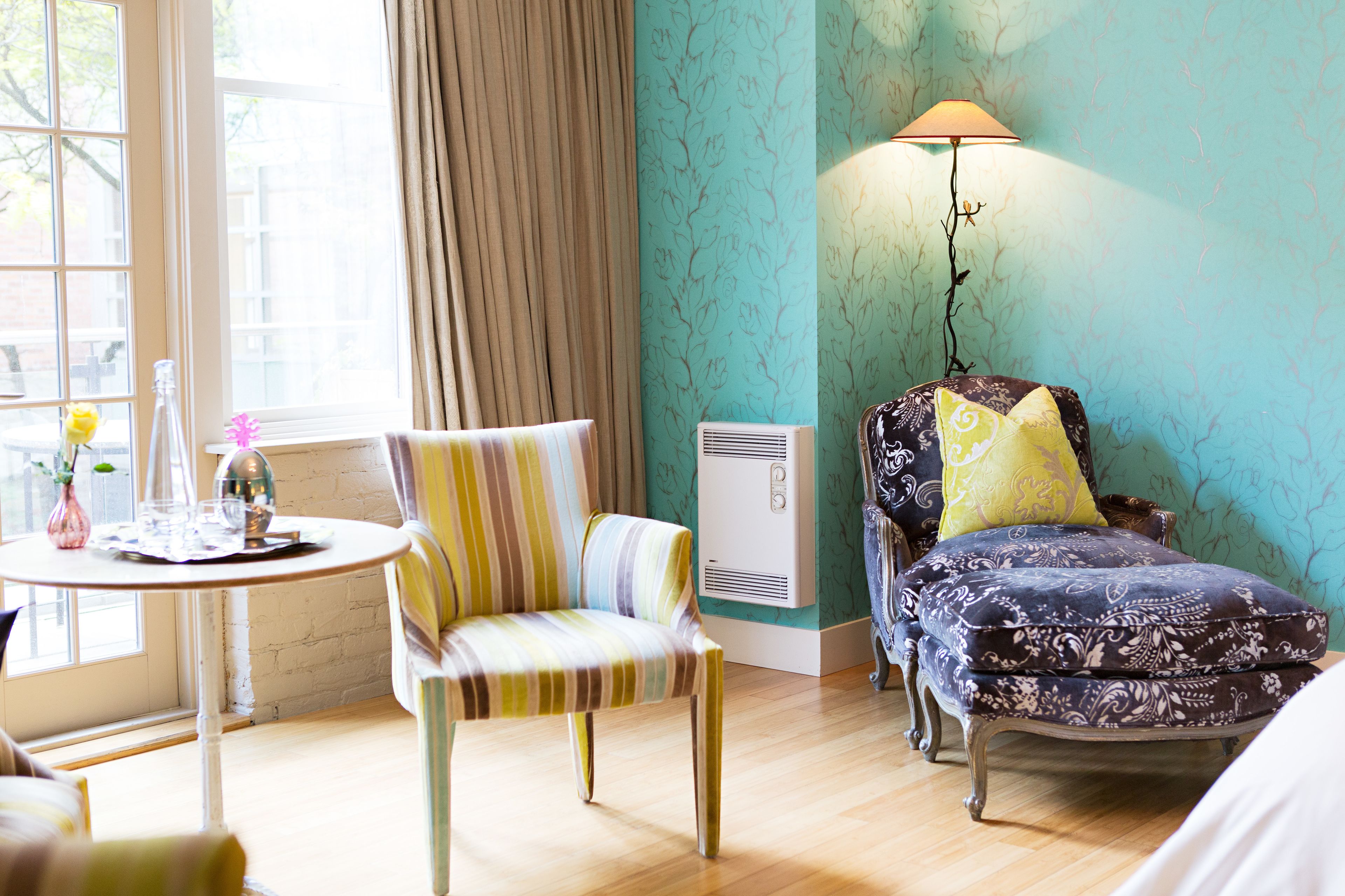 Una habitación con un sillón a rayas, una mesa redonda con un florero y una botella, un sillón estampado con un cojín amarillo y una lámpara de pie.