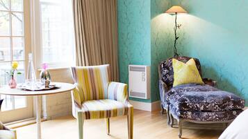 Una habitación con un sillón a rayas, una mesa redonda con un florero y una botella, un sillón estampado con un cojín amarillo y una lámpara de pie.