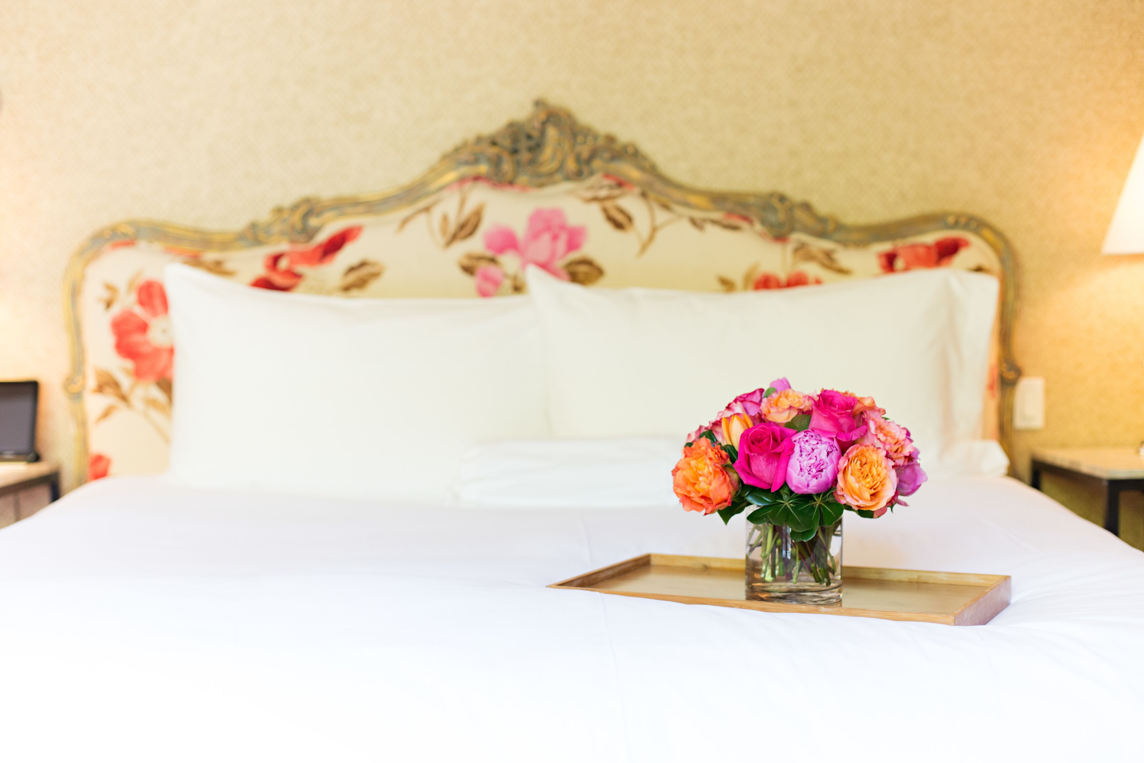 Una cama con cabecera floral, un jarrón con flores sobre una bandeja y una lámpara en la mesita de noche.