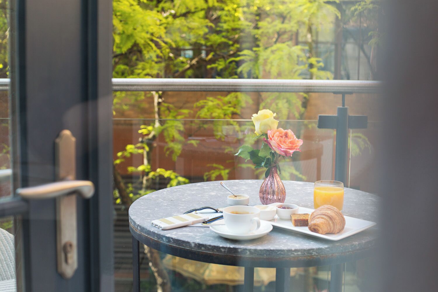 A round table with a glass top, a vase with flowers, a croissant, a glass of orange juice, and a cup of coffee.