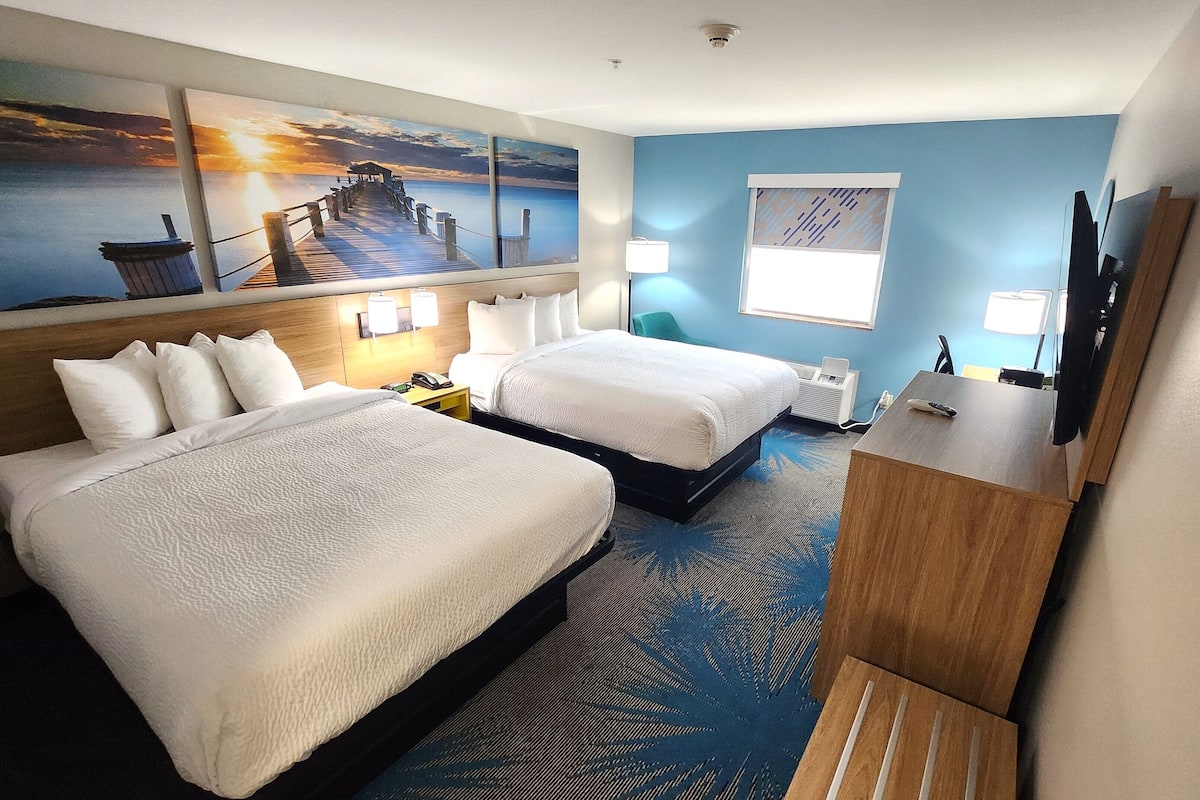 Habitación de hotel con dos camas, un escritorio, un televisor y una obra de arte que representa un atardecer sobre el agua.