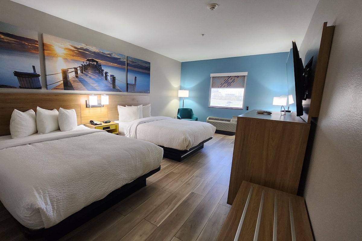 Habitación de hotel con dos camas, cabecera de madera, un escritorio con silla, televisor y una obra de arte en la pared.