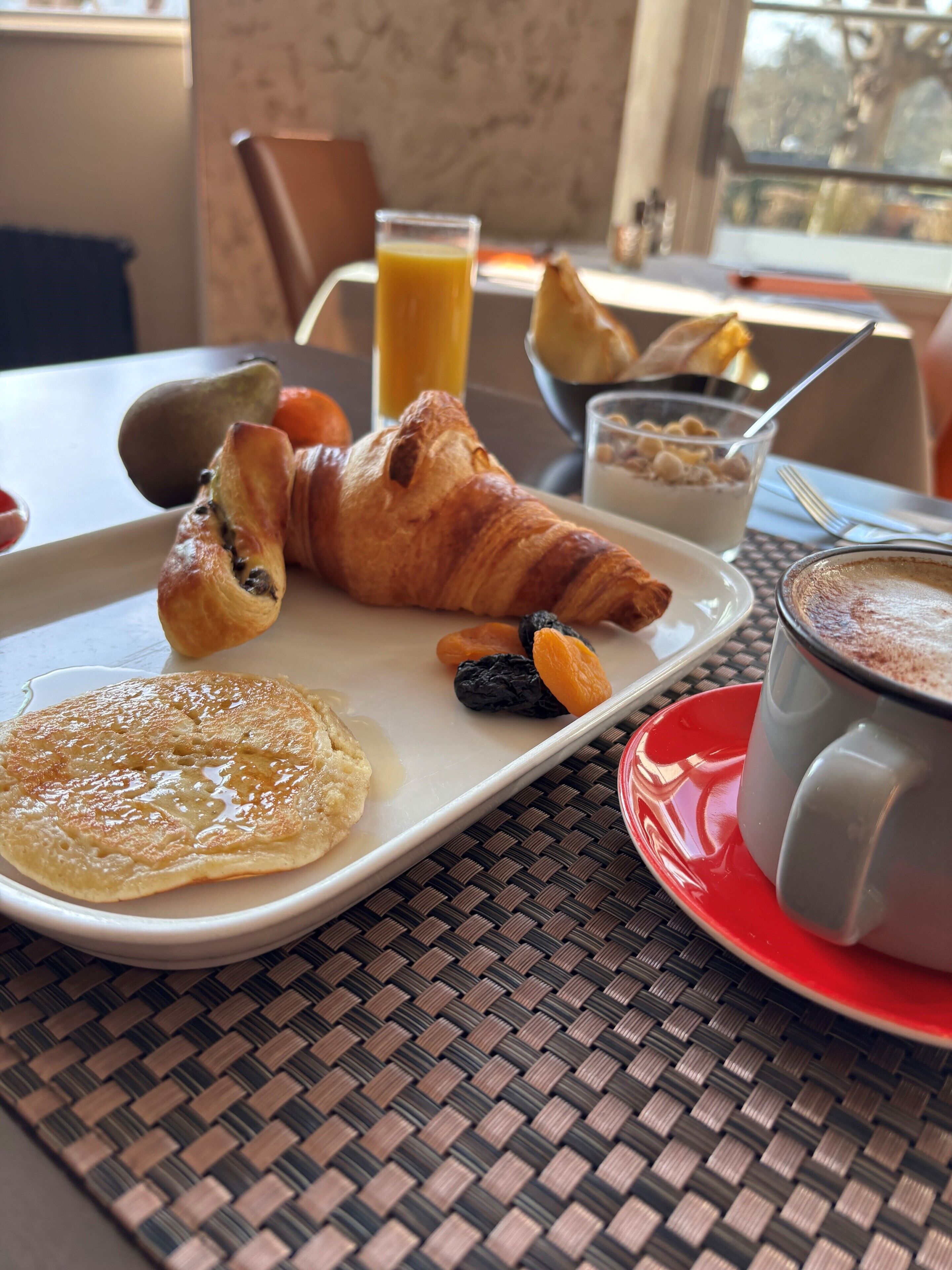 Un plato de desayuno con cruasanes, un panqueque y frutas secas, junto con un vaso de jugo de naranja y una taza de café.