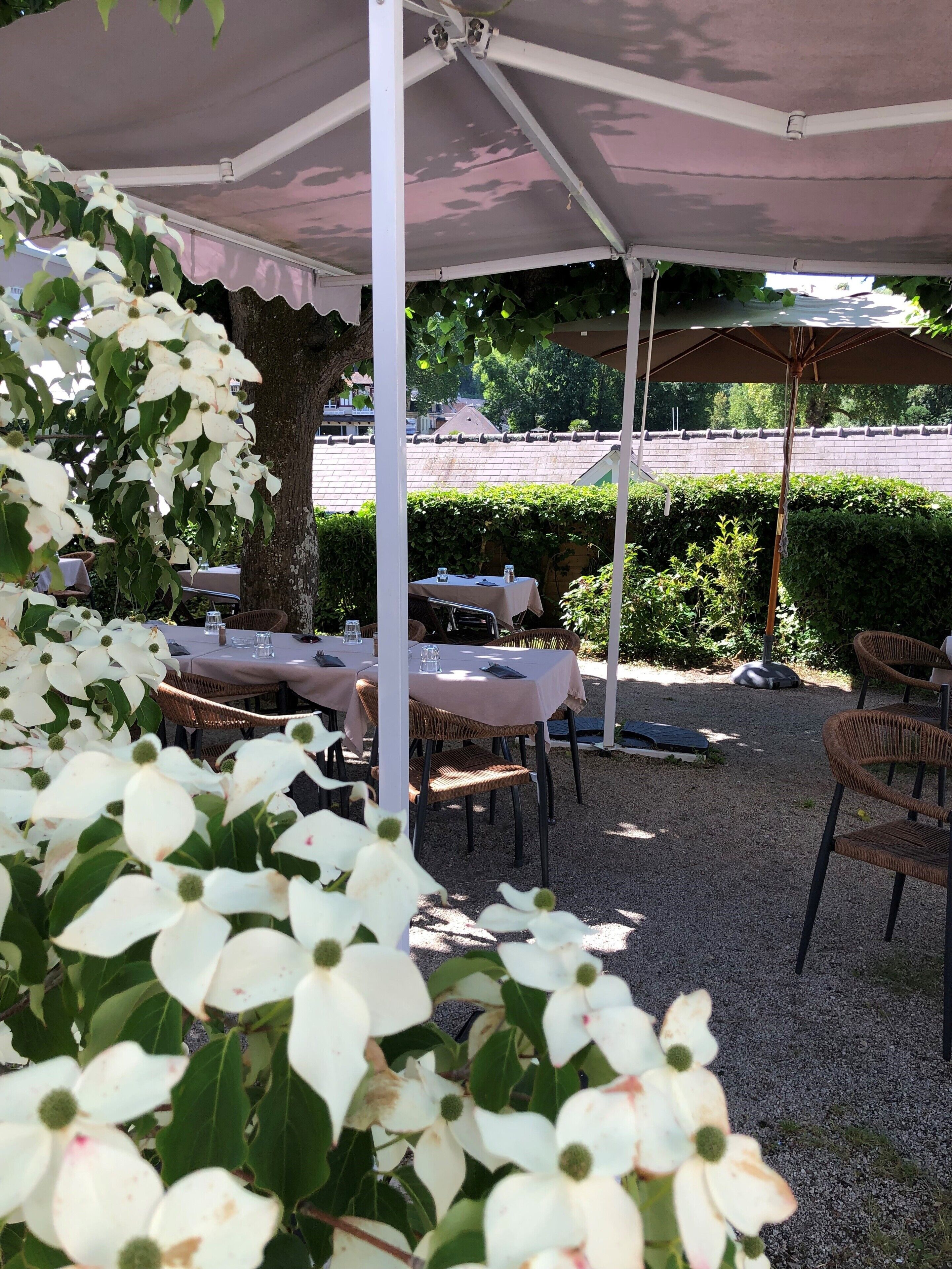 Eine Außenpatio mit Tischen und Stühlen unter Sonnenschirmen, umgeben von Pflanzen und einer Hecke.