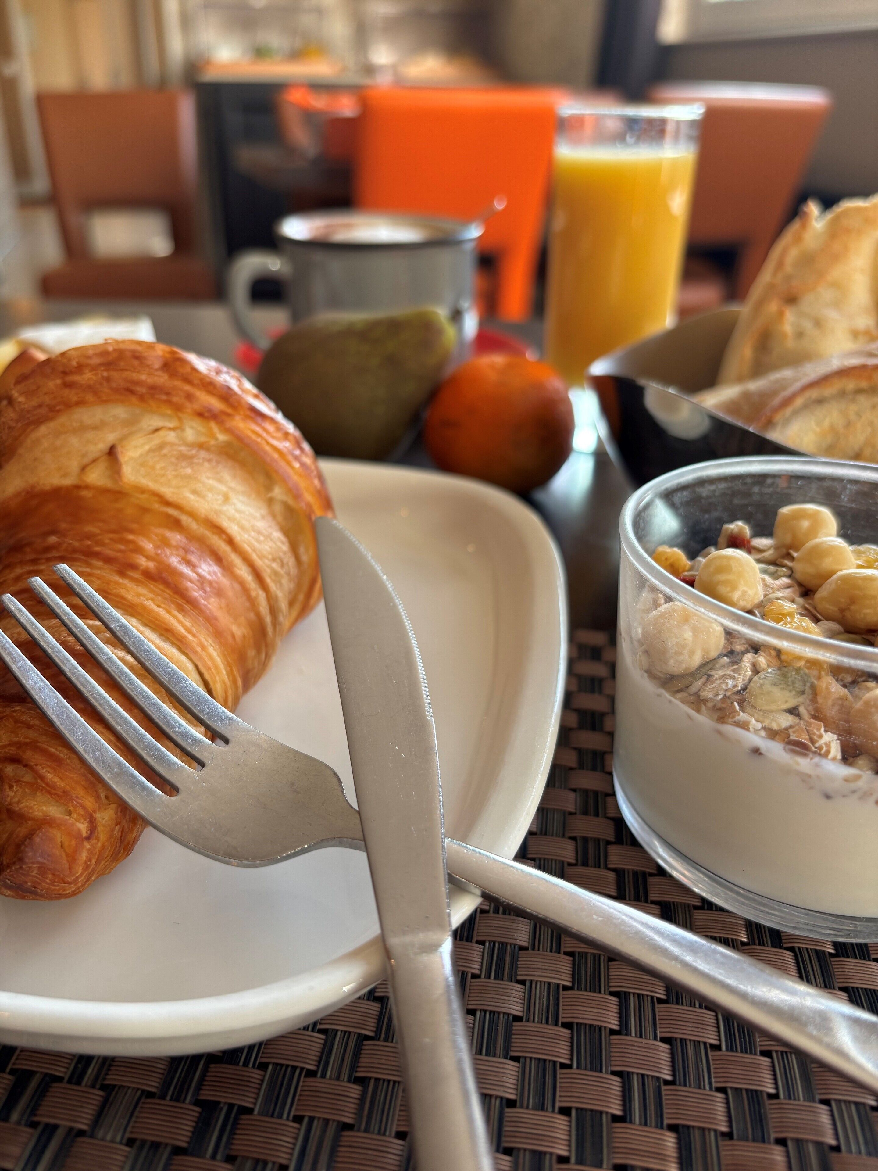 Un desayuno con un croissant, un vaso de jugo de naranja, un tazón de cereal y una taza de metal.