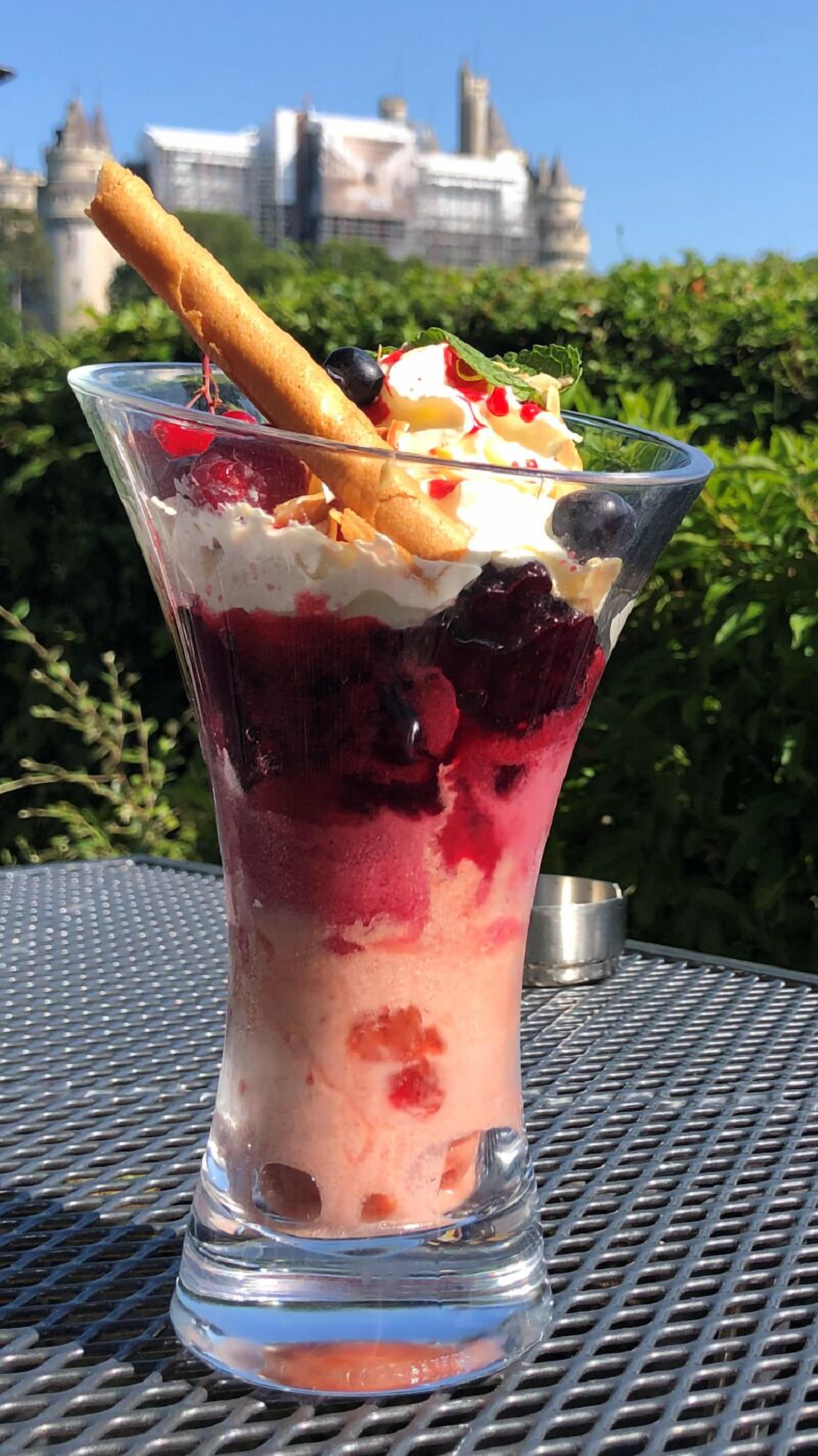 A dessert in a clear glass with berries, whipped cream, and a wafer stick, set on a metal table with greenery and a building in the background.