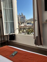 Habitación de hotel con cama, ventana con cortinas, vista a un castillo y barandilla de balcón.