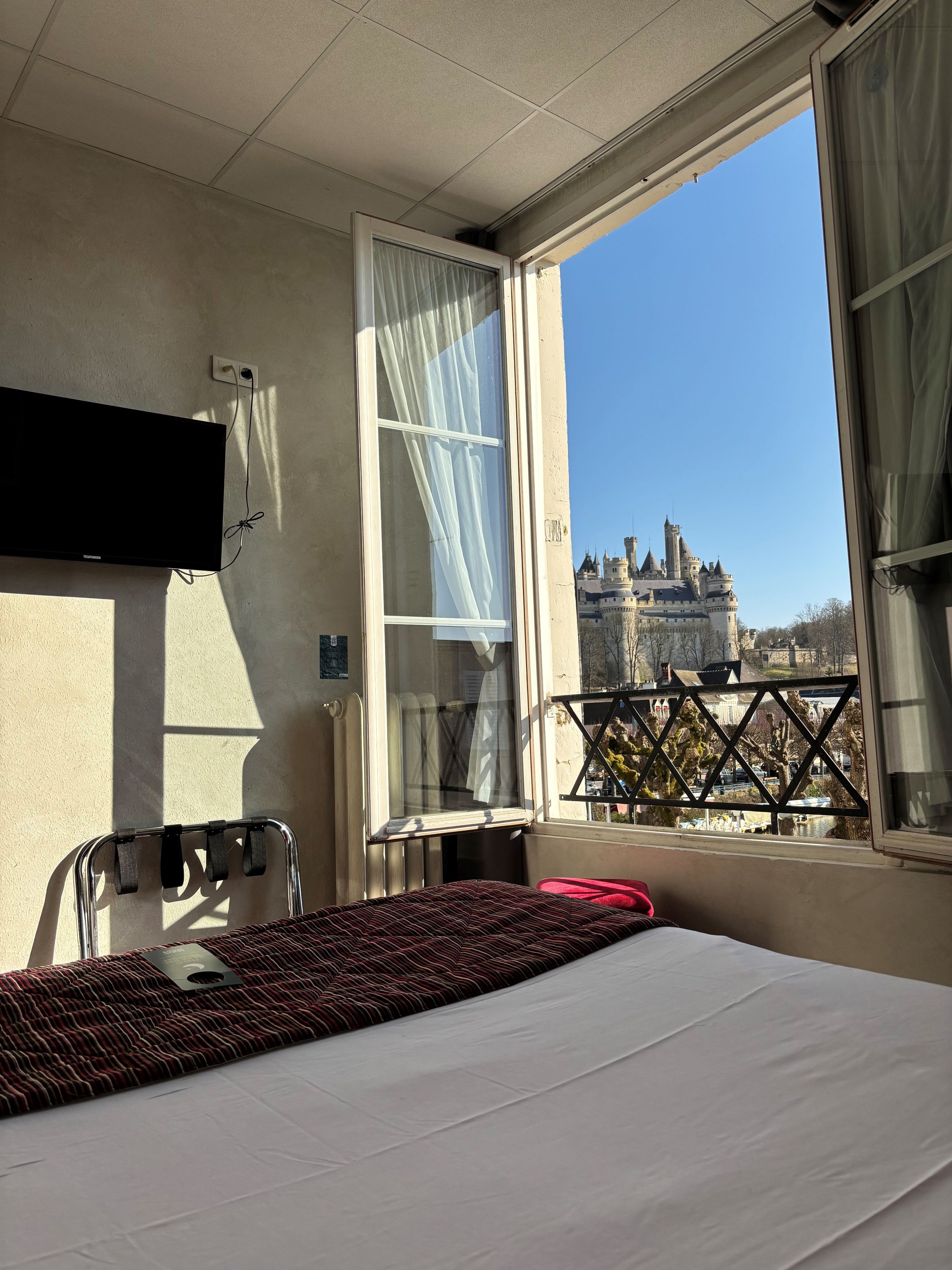 A hotel room with a bed, a TV, a window with curtains, and a view of a castle.