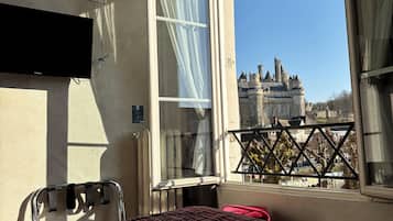 A hotel room with a bed, a TV, a window with curtains, and a view of a castle.