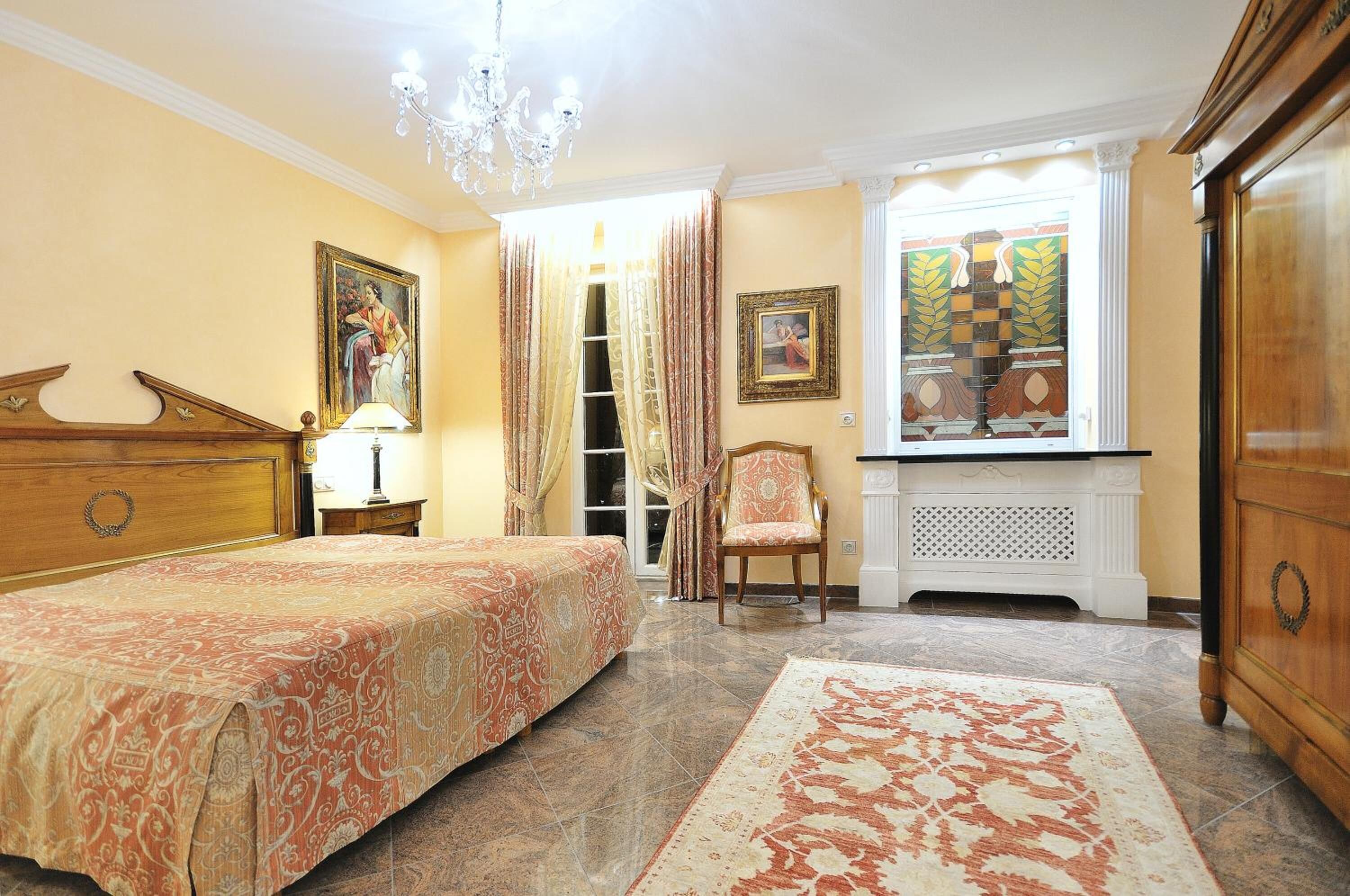 Un dormitorio con cama, una silla, cómoda, ventana con cortinas y candelabro.