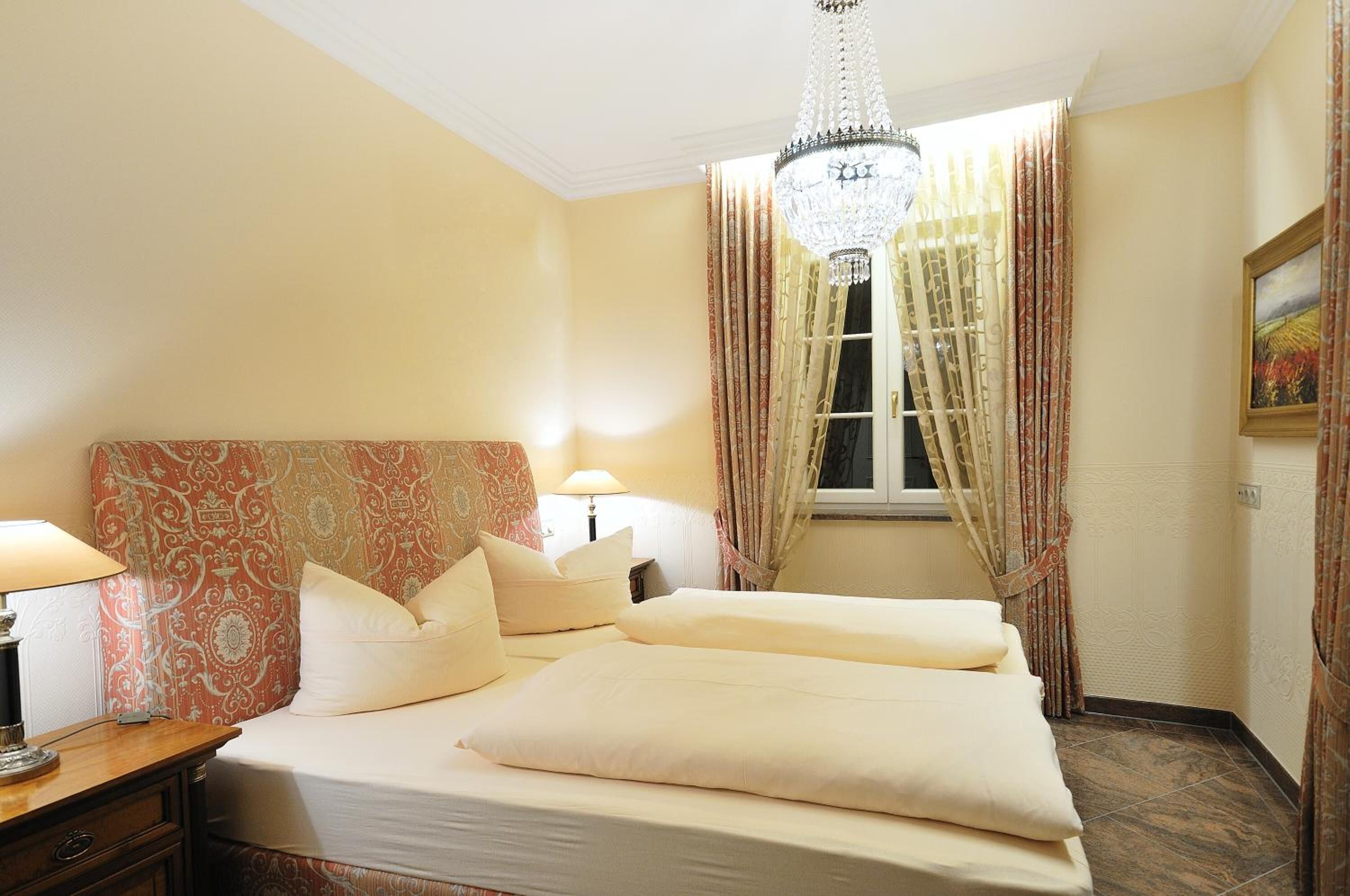 Una habitación de hotel con una cama grande, dos lámparas de noche, una araña y cortinas estampadas.