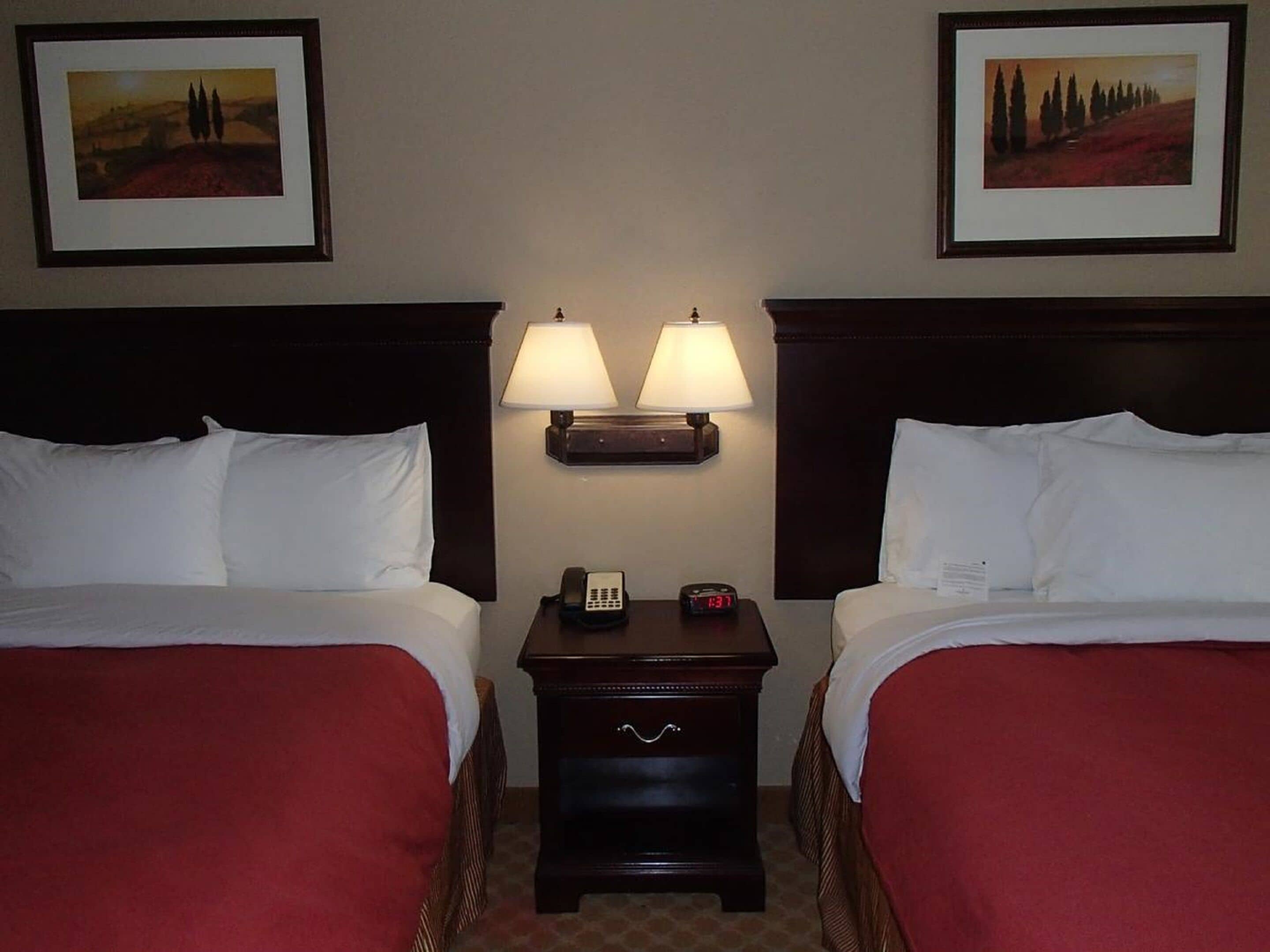 Habitación de hotel con dos camas, cada una con sábanas blancas y colchas rojas. Entre las camas hay dos lámparas de pared, y una mesita de noche con un teléfono y un despertador.