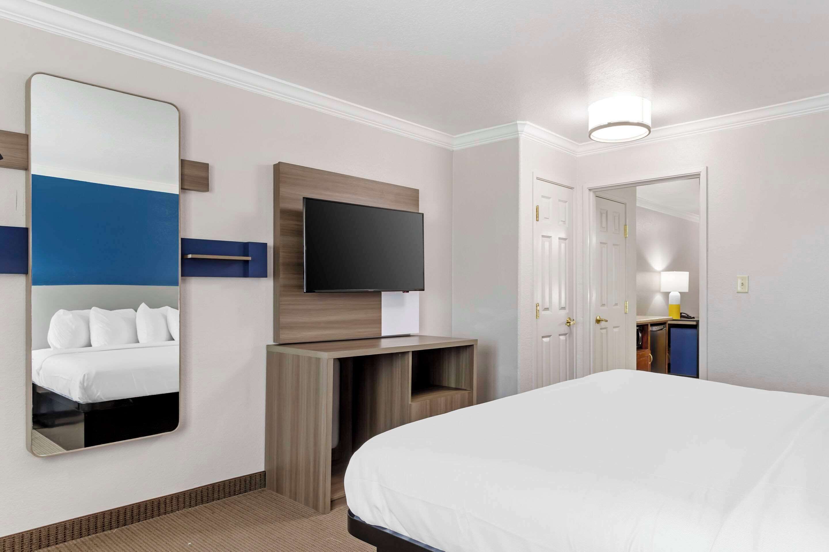 A hotel room with a large bed, a flat-screen TV mounted on a wooden panel, a mirror reflecting the bed, a door leading to a bathroom, and a small desk with a lamp.