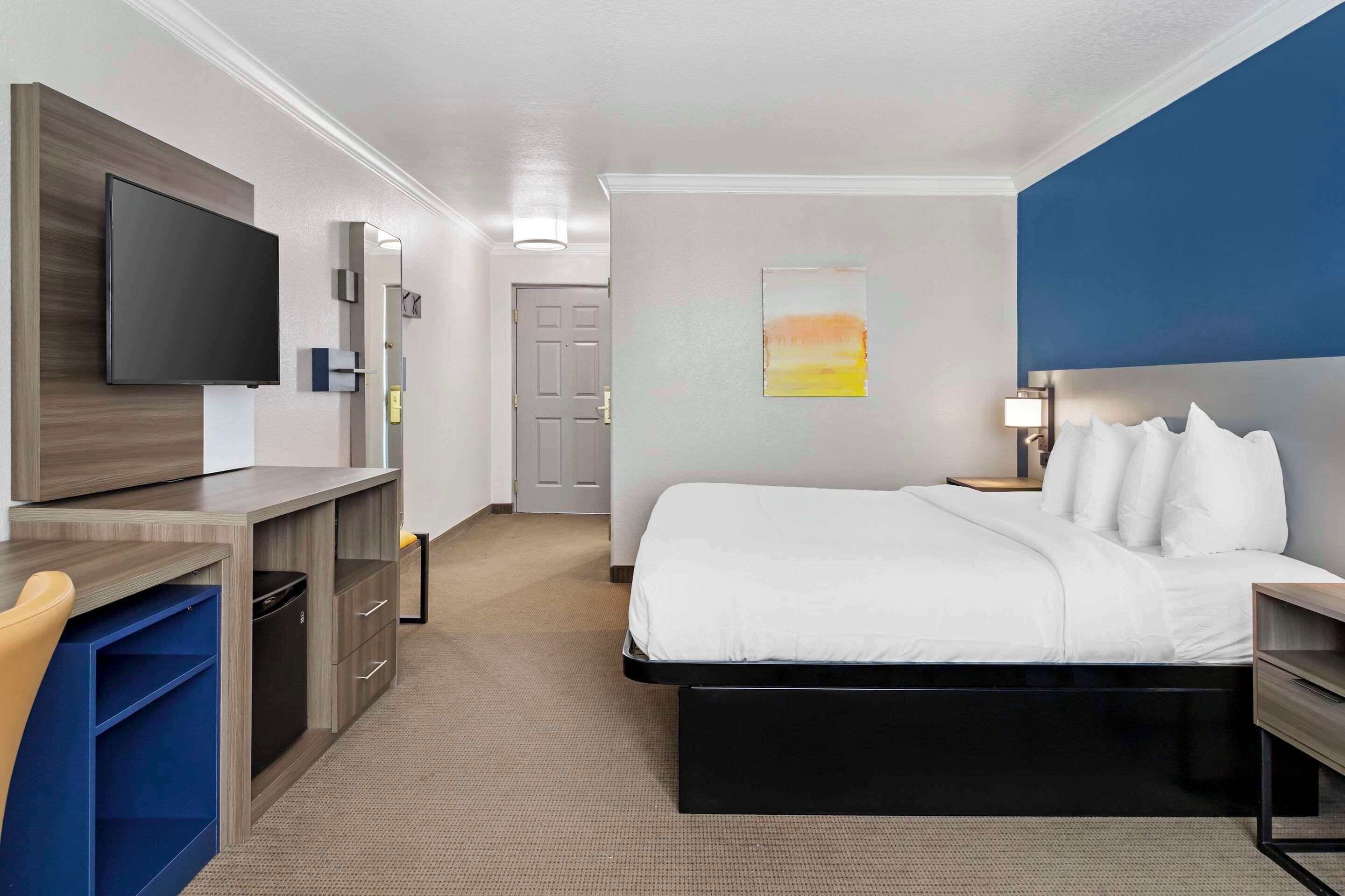A hotel room with a bed, a desk with a chair, a television, a nightstand, and a painting on the wall.