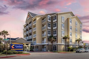 A multi-story hotel with balconies, a prominent Comfort Inn & Suites sign, and a street view with palm trees and parked cars.