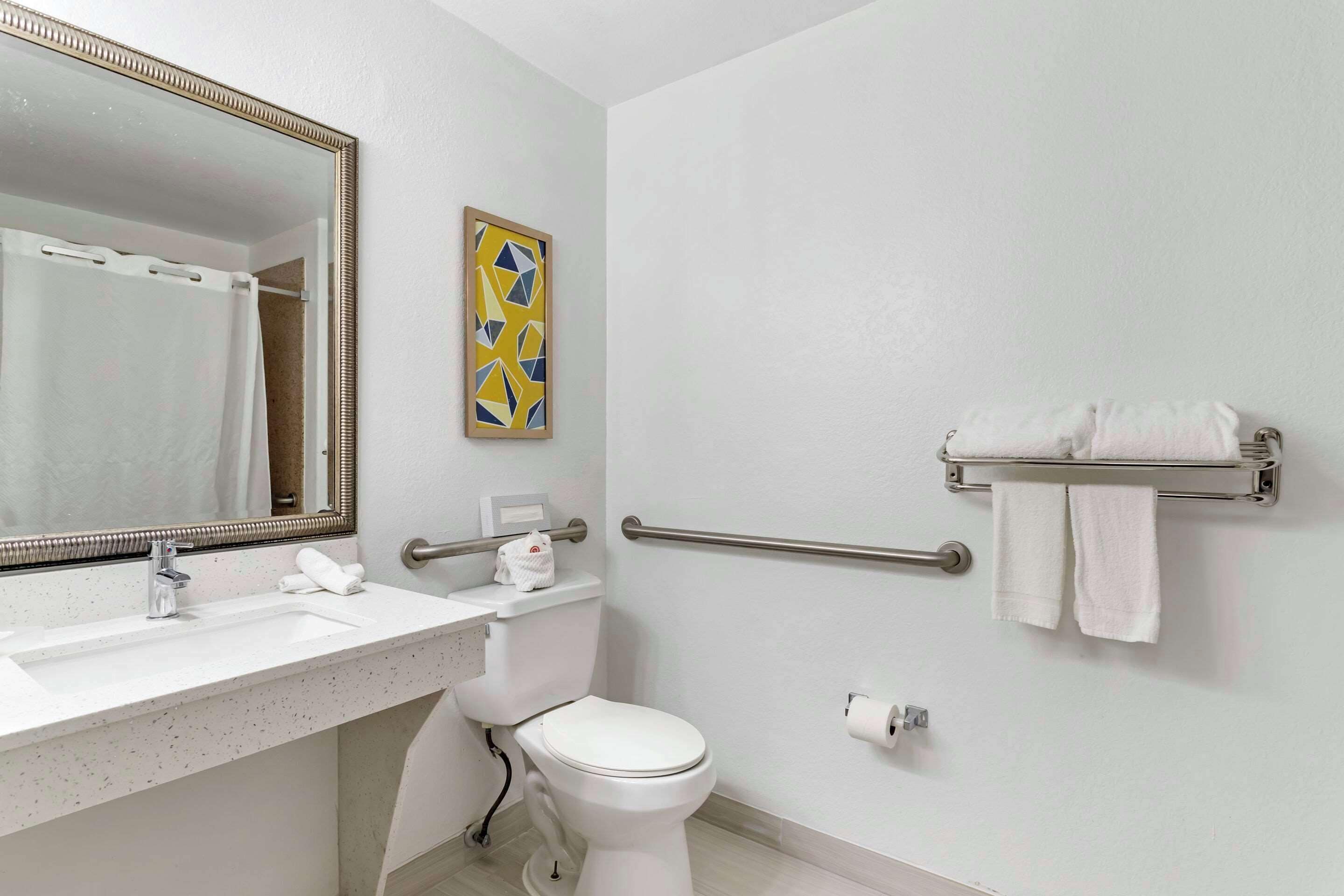 Un baño con lavamanos blanco, inodoro, espejo, toallero y dispensador de papel higiénico.