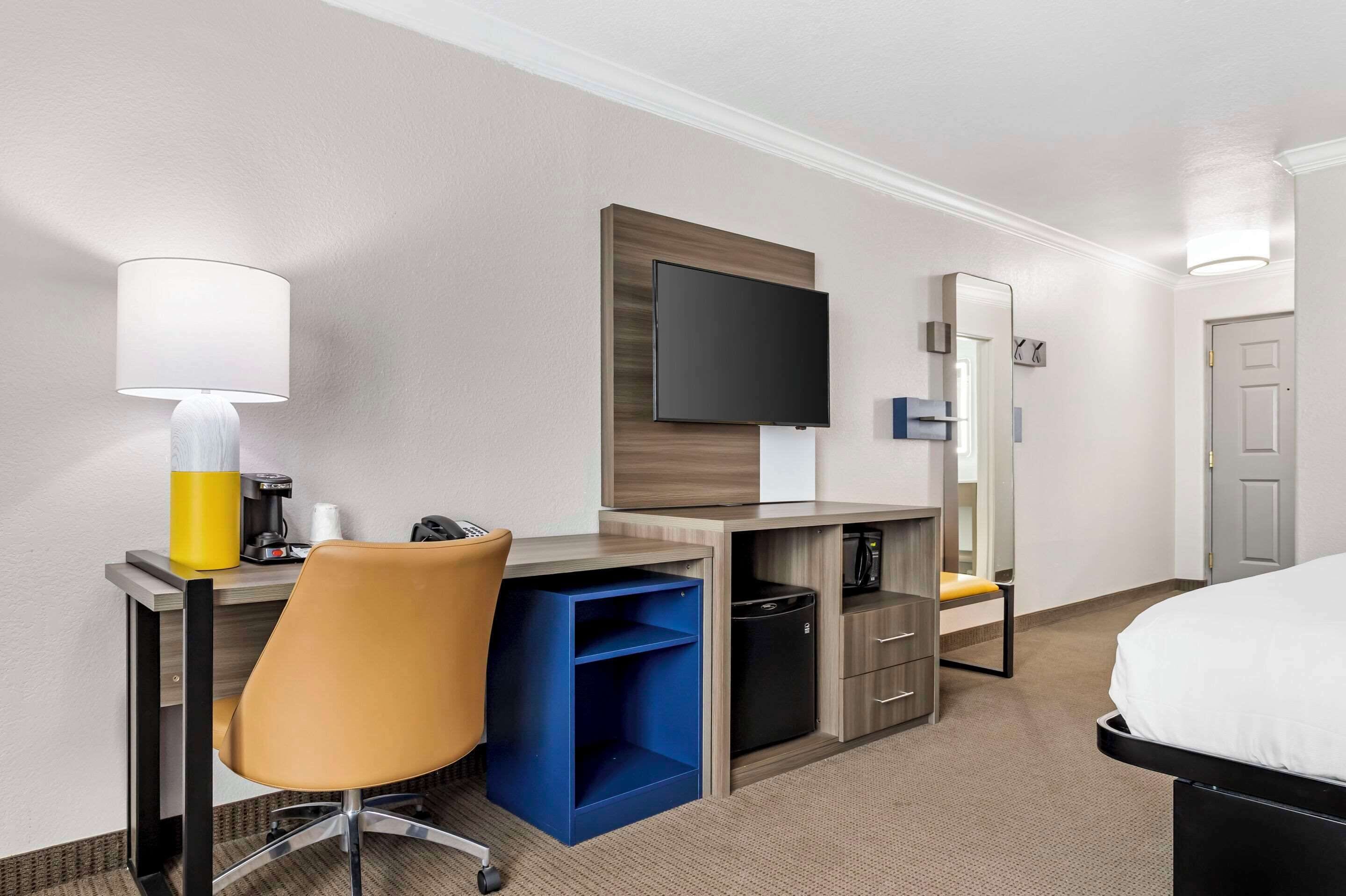 A hotel room with a bed, a desk with a chair, a TV, a coffee machine, and a small blue storage unit.