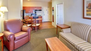 A living room with a leather armchair, a striped sofa, and a wooden coffee table. There is a kitchen in the background with wooden cabinets and a refrigerator. - Candlewood Suites Ft Stockton by IHG (Fort Stockton)