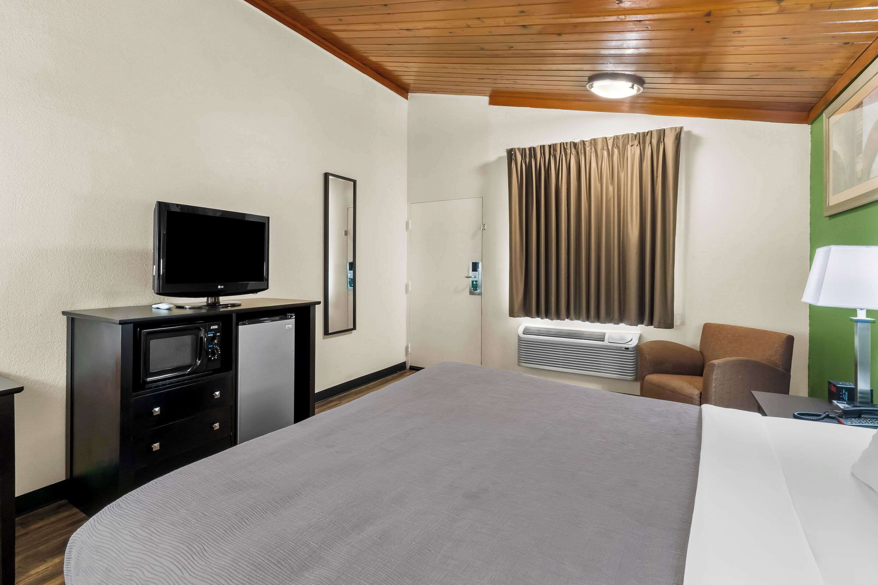 Une chambre d’hôtel comprenant un lit, un bureau avec une lampe, une télévision, un mini-réfrigérateur, un four à micro-ondes, une chaise et une fenêtre avec des rideaux.