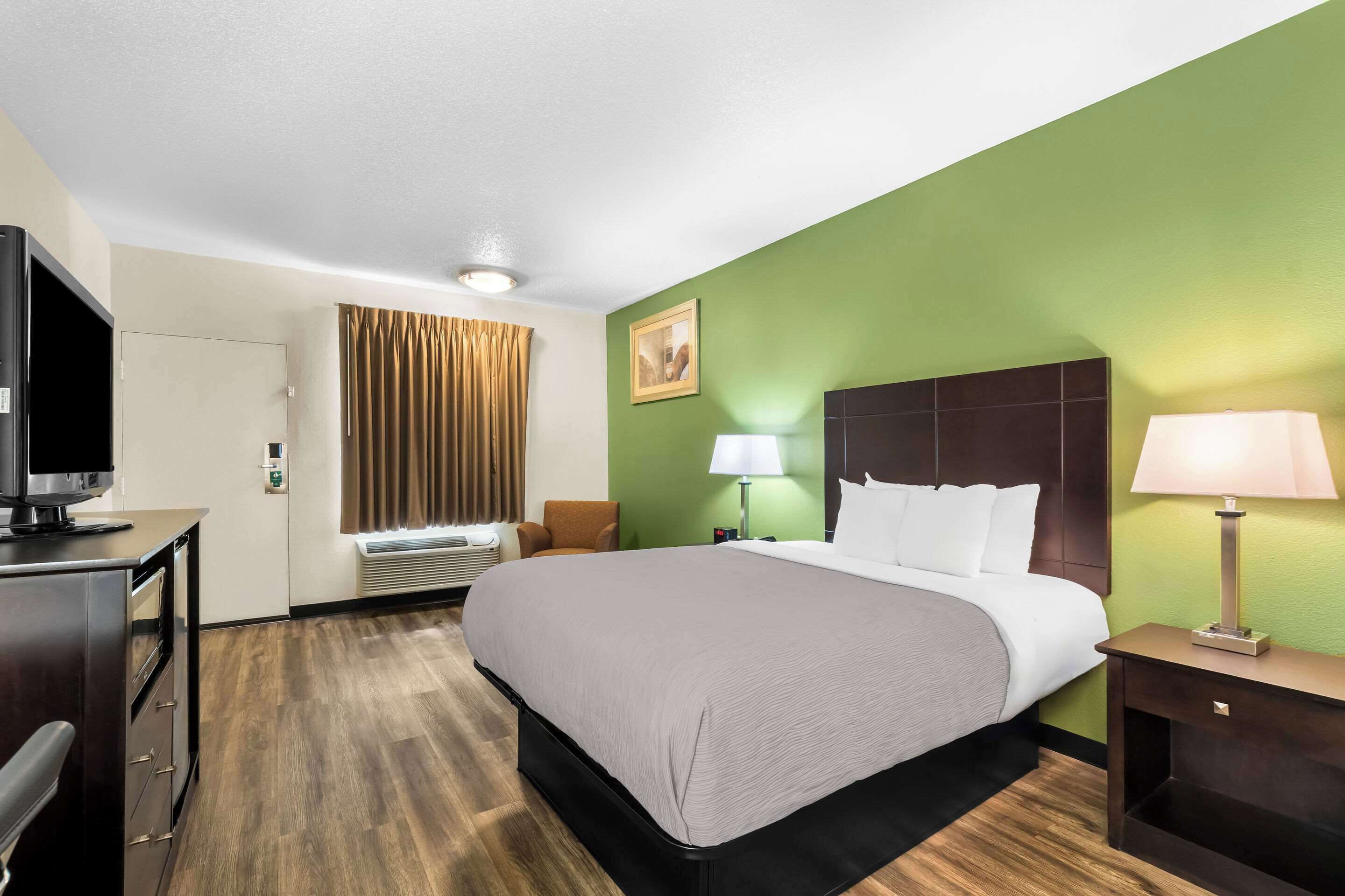 A hotel room with a bed, a desk with a television, a chair, a lamp, and a picture on the wall.