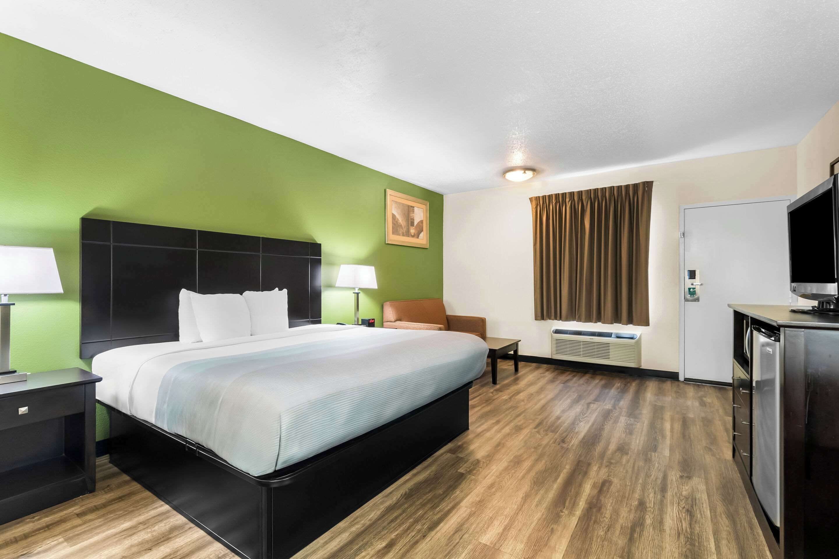 Une chambre d’hôtel avec un lit, un bureau, une chaise, une lampe, un tableau au mur et une fenêtre avec des rideaux.