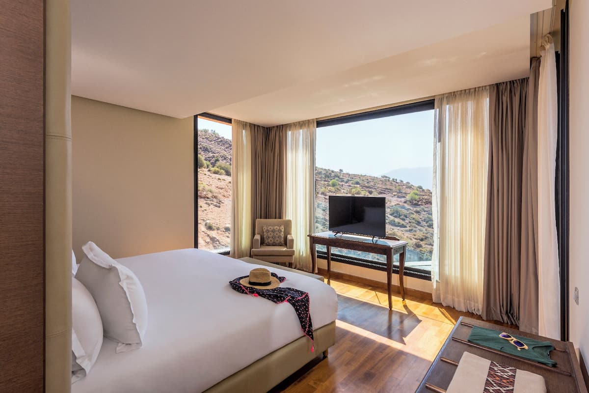 Habitación de hotel con una cama grande, un televisor sobre una mesa de madera y vistas a un paisaje montañoso.