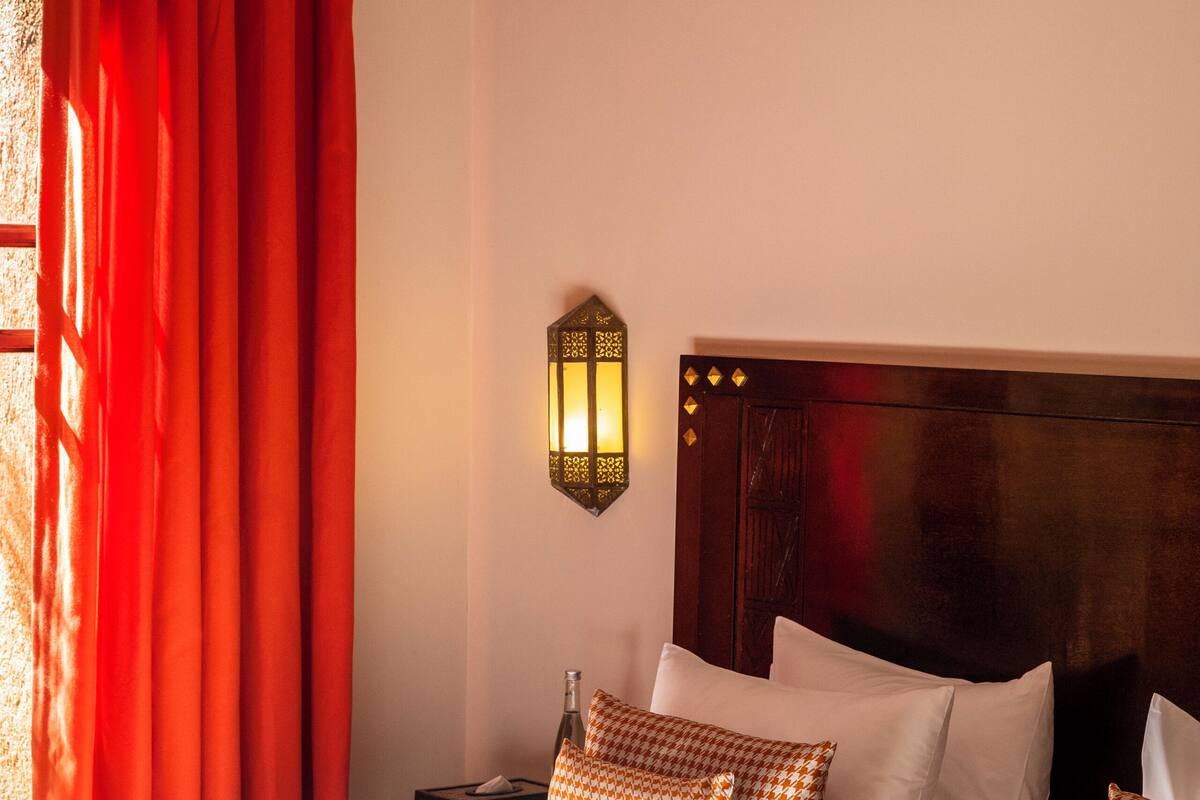 Una habitación de hotel con una cama grande, cortinas rojas, una lámpara de pared y una colcha estampada.