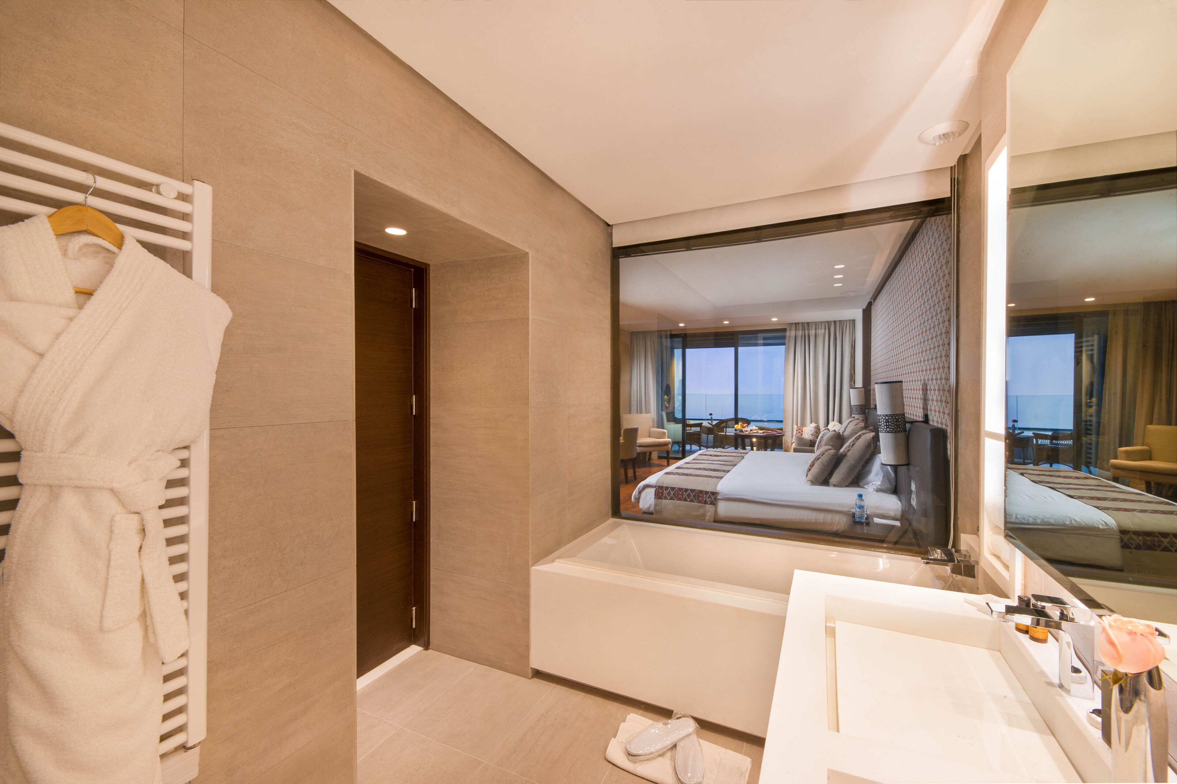 A modern hotel room with a large bed, a bathtub, a vanity with a mirror, and a view of the city through a large window.