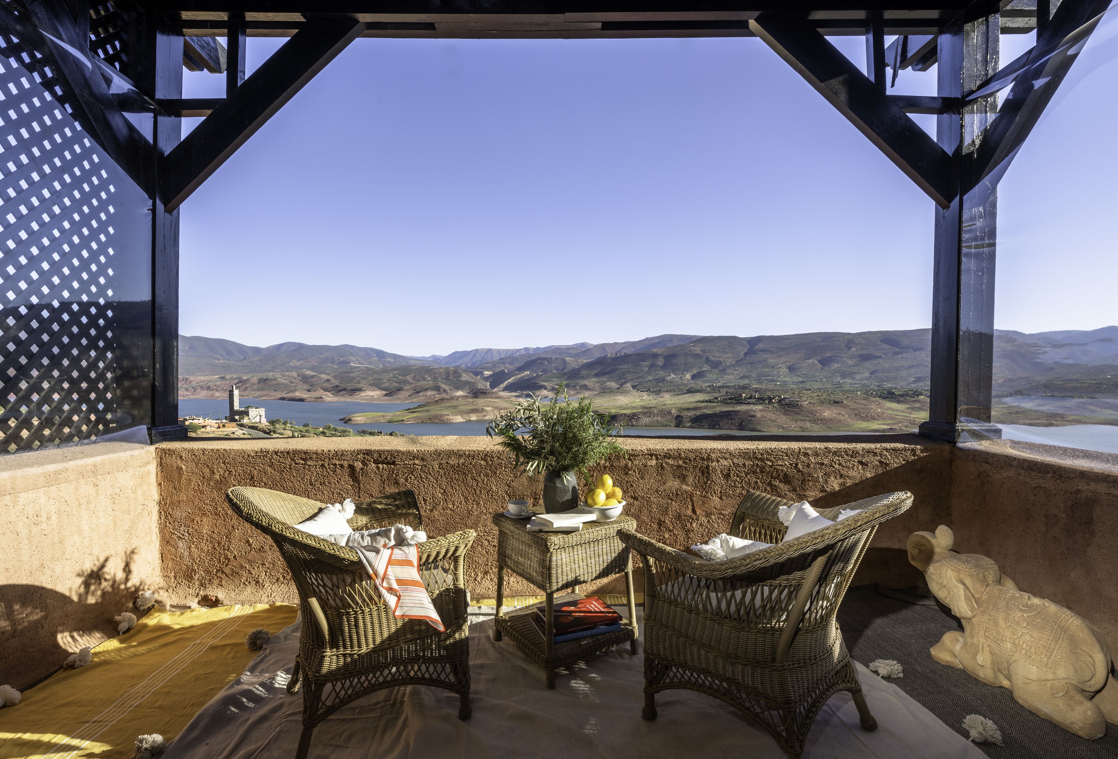 Une terrasse meublée de mobilier en osier, une table ornée d’un vase et de citrons, et une vue panoramique sur des collines et un lac.