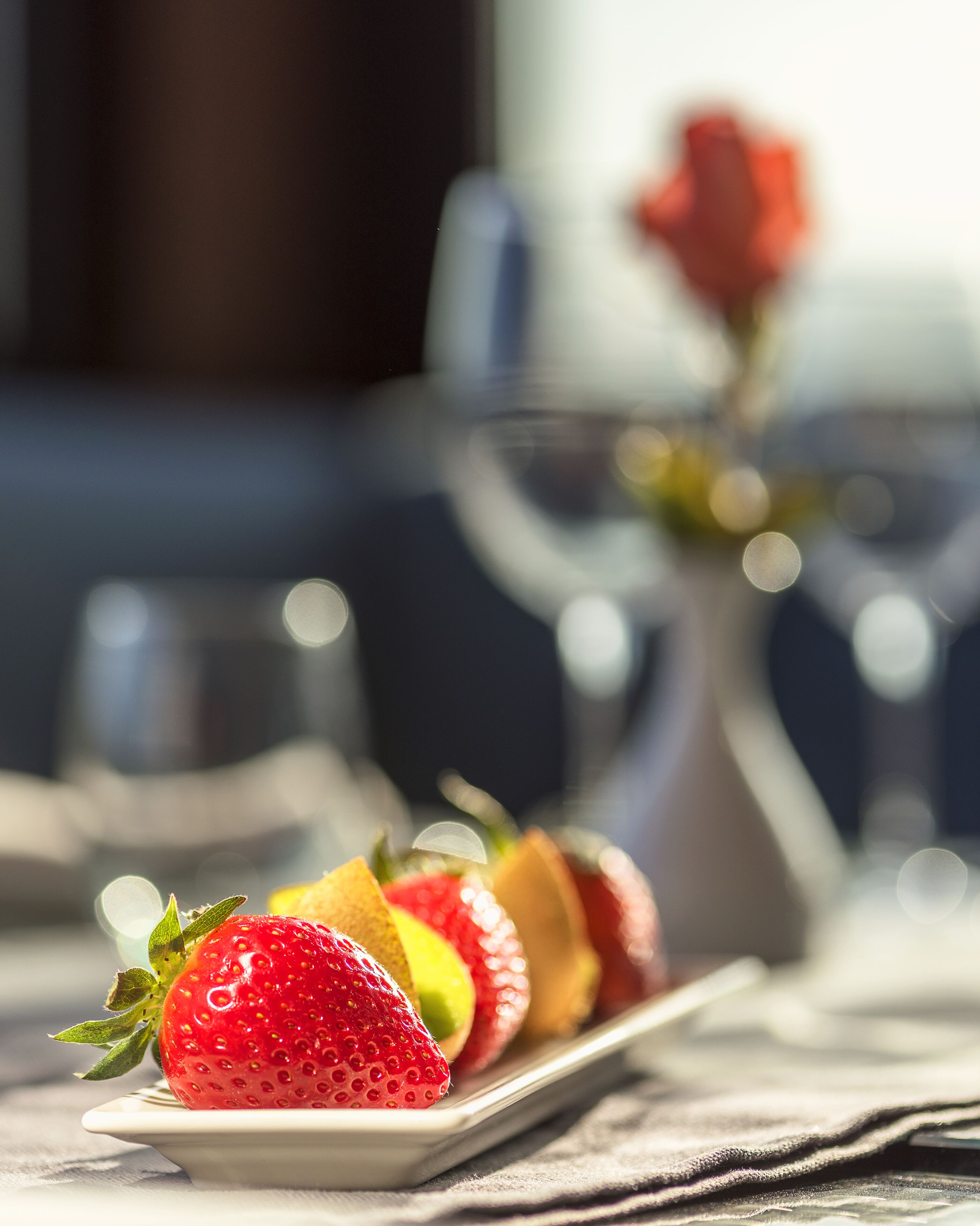 Un plato de fruta que incluye fresas y una rebanada de kiwi sobre una mesa, con un fondo desenfocado que muestra un jarrón con una rosa.