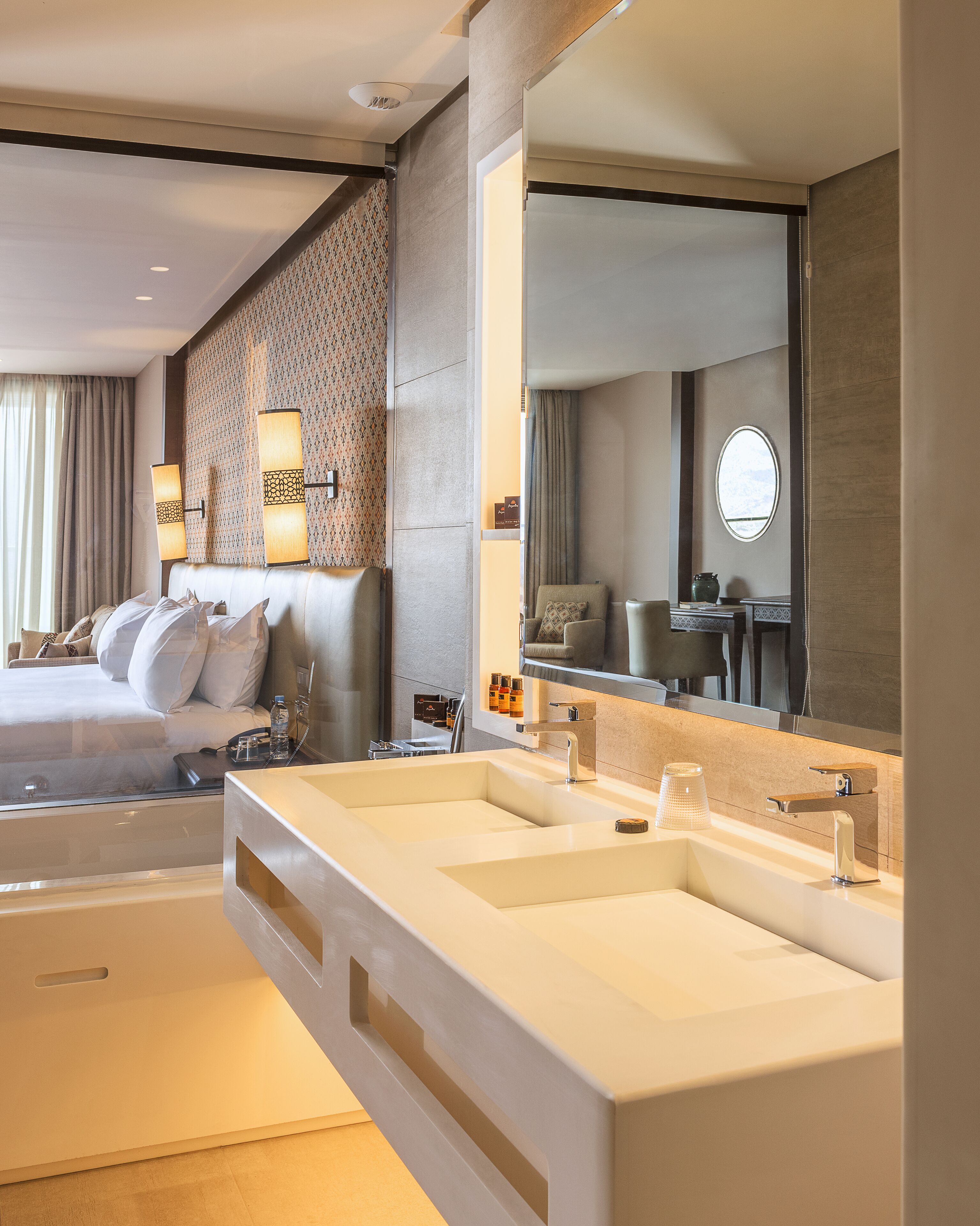 A modern hotel room with a large bed, a separate sitting area, a large mirror, and a dual sink vanity.