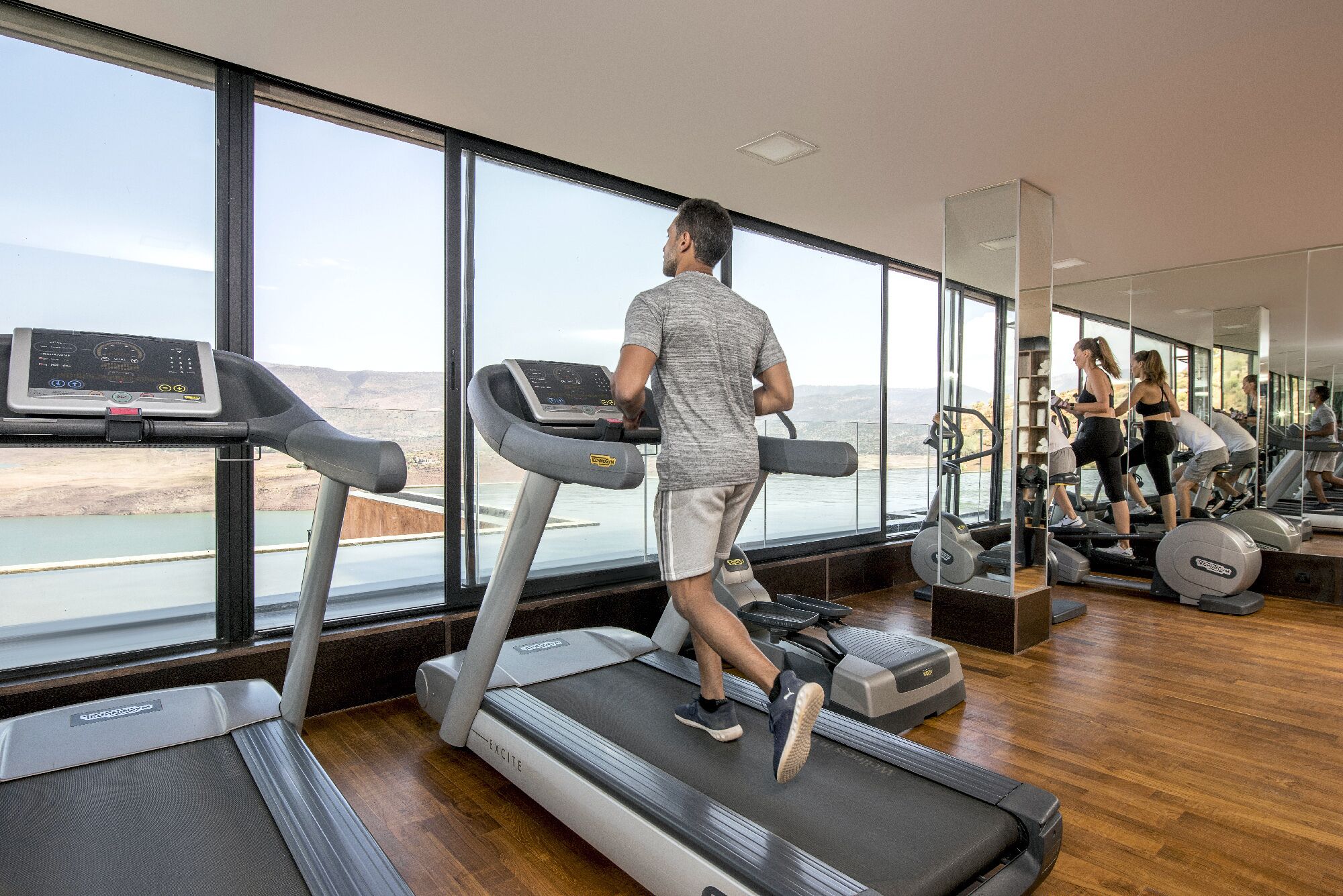 A gym with treadmills, exercise equipment, and large windows offering a view of a body of water and landscape.