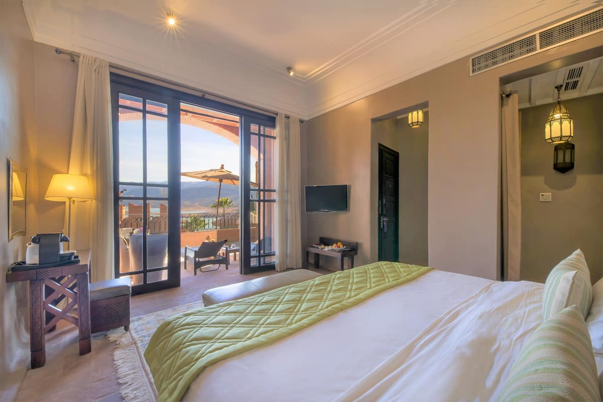 Una habitación de hotel con cama, un escritorio con lámpara, televisión y balcón con sombrilla.