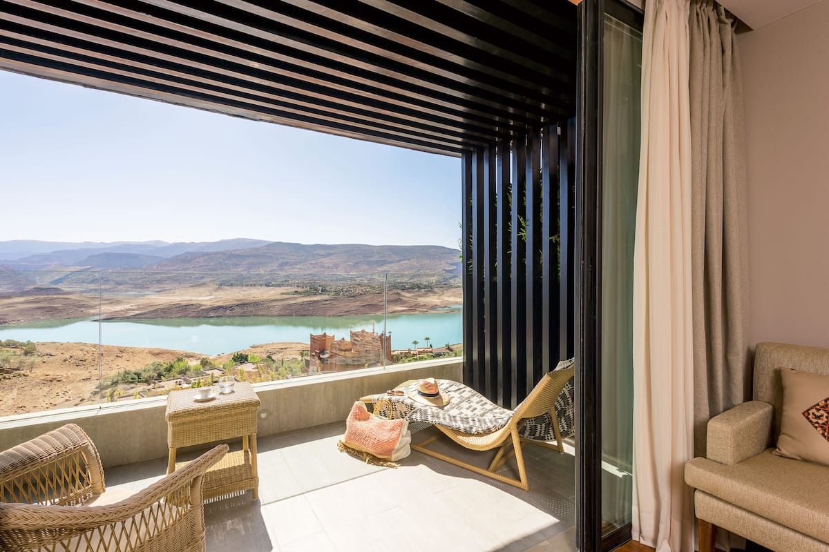 Un balcón con muebles de mimbre, una mesa con bebidas y vistas a un lago y montañas.