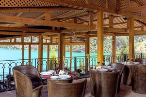 An outdoor dining area with wooden beams and a view of a lake. Tables are set with plates and glasses.