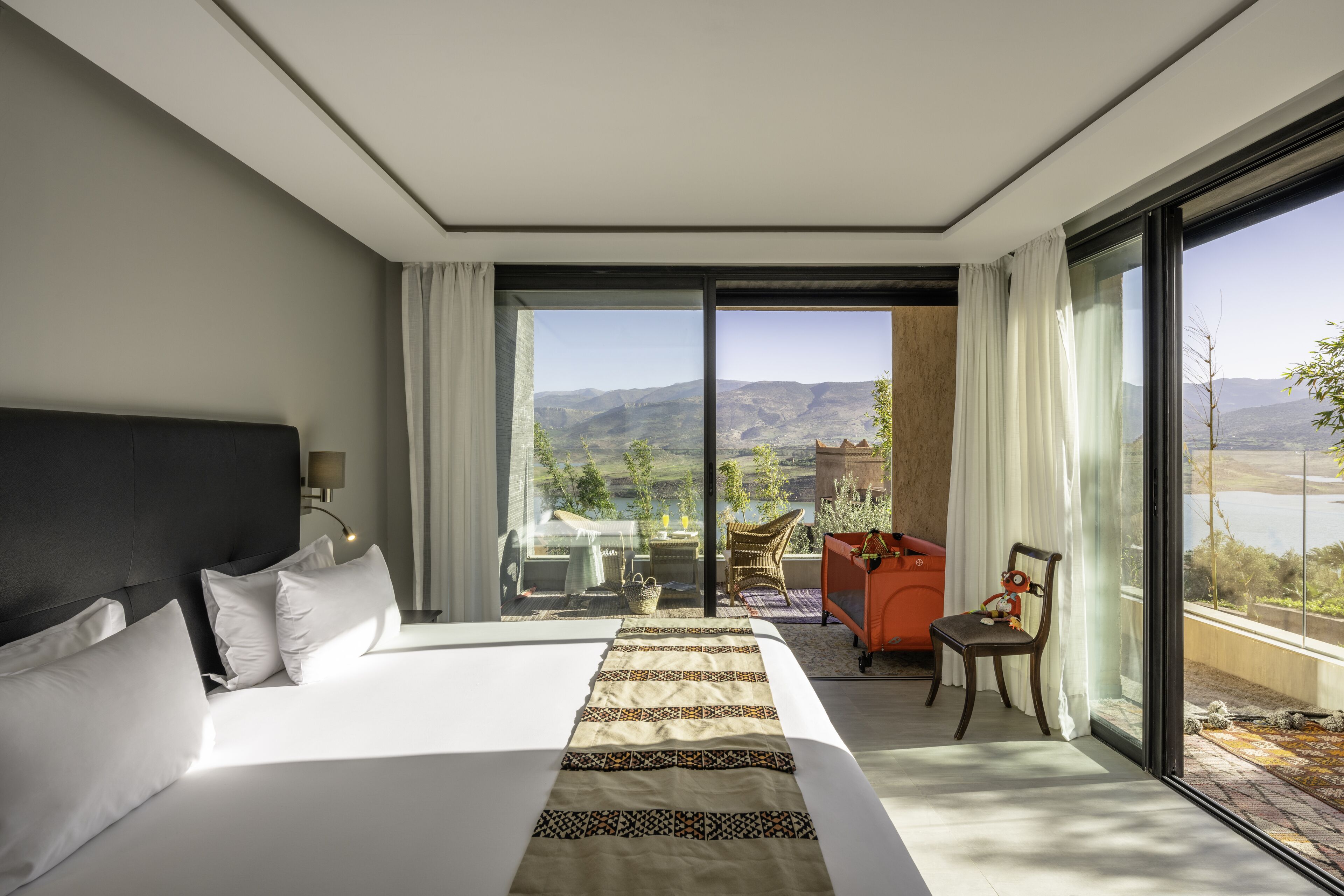 A modern hotel room with a large bed, a view of the mountains, and a balcony with outdoor furniture.
