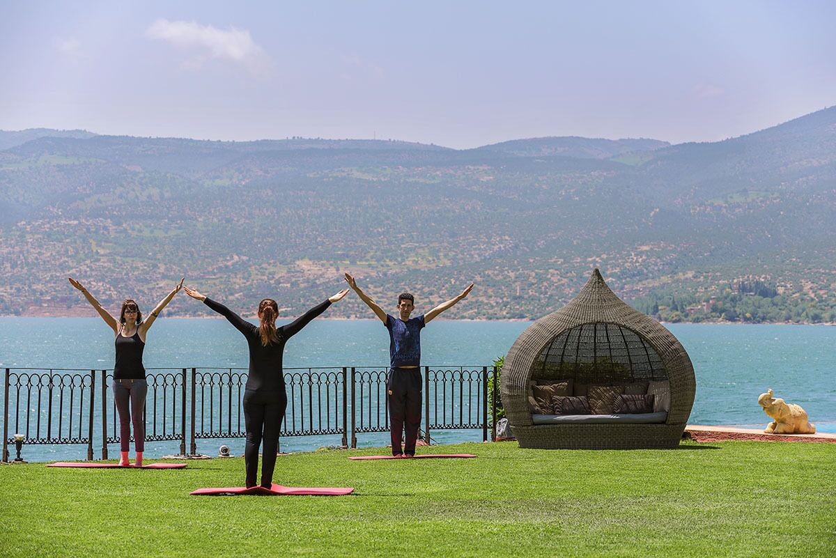 Drei Personen üben Yoga auf einer Wiese mit Blick auf einen See und Hügel im Hintergrund.