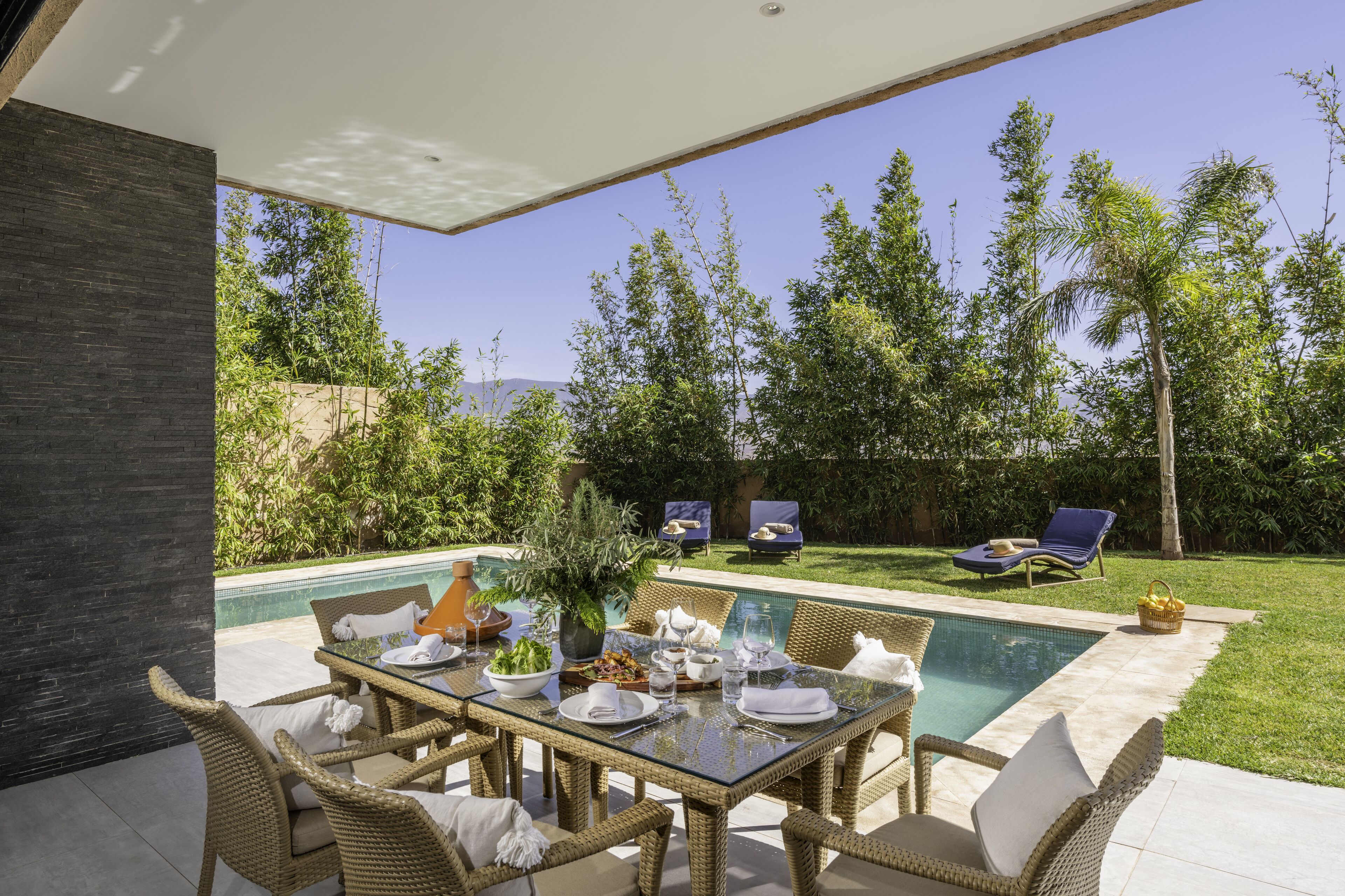 Un área de comedor exterior moderna con una mesa de vidrio, sillas de mimbre y una piscina al fondo.