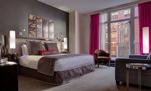 Quarto de hotel moderno com uma cama grande, uma cadeira, uma mesinha e uma janela com cortinas.