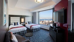 Habitación de hotel con dos camas, un sofá, un televisor y un amplio ventanal con vista a la ciudad.