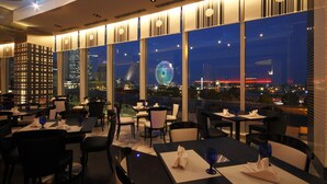 Un restaurante con mesas y sillas, ventanas amplias y vistas a un paisaje urbano nocturno.