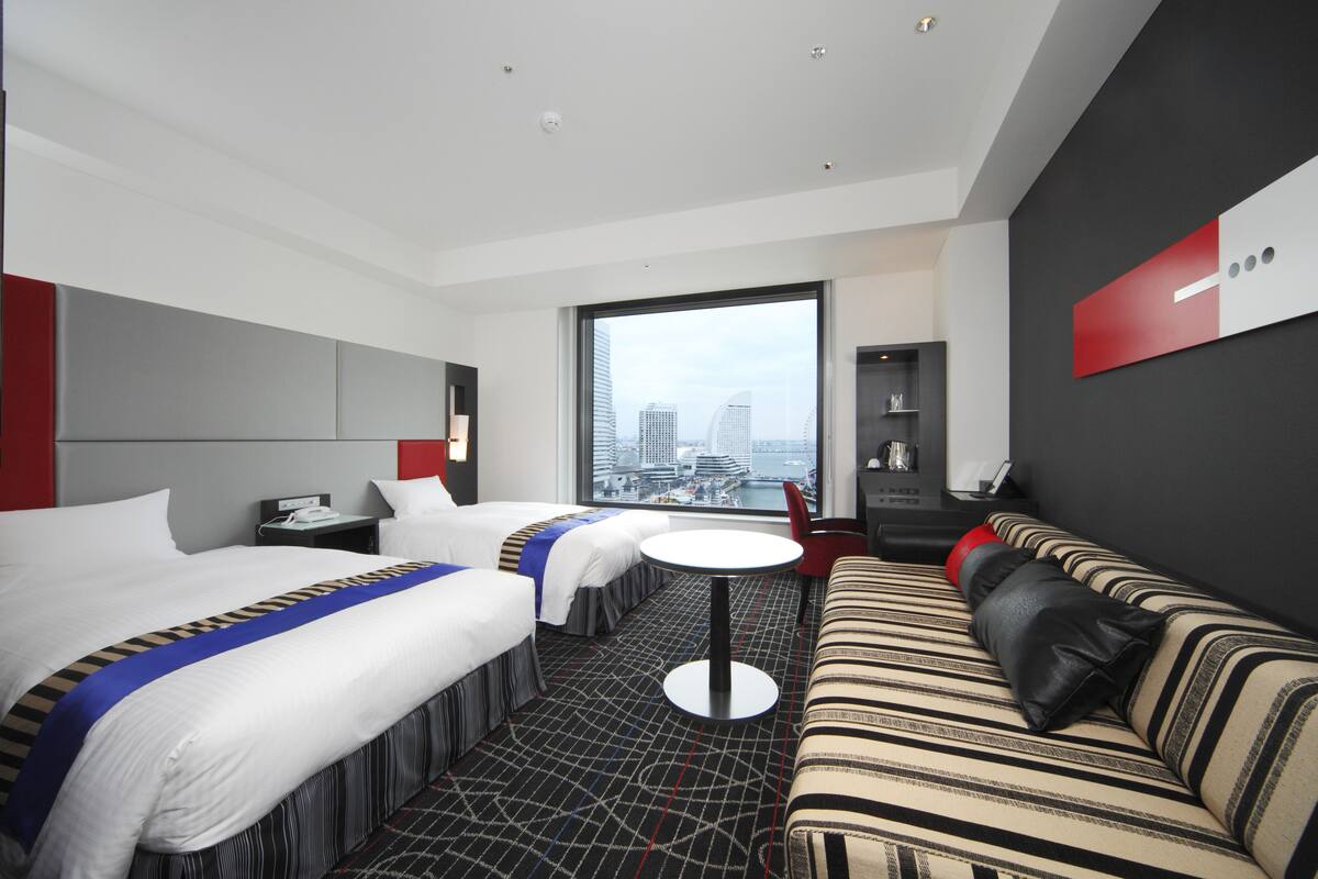 A hotel room with two beds, a sofa, a small table, and a large window with a city view.